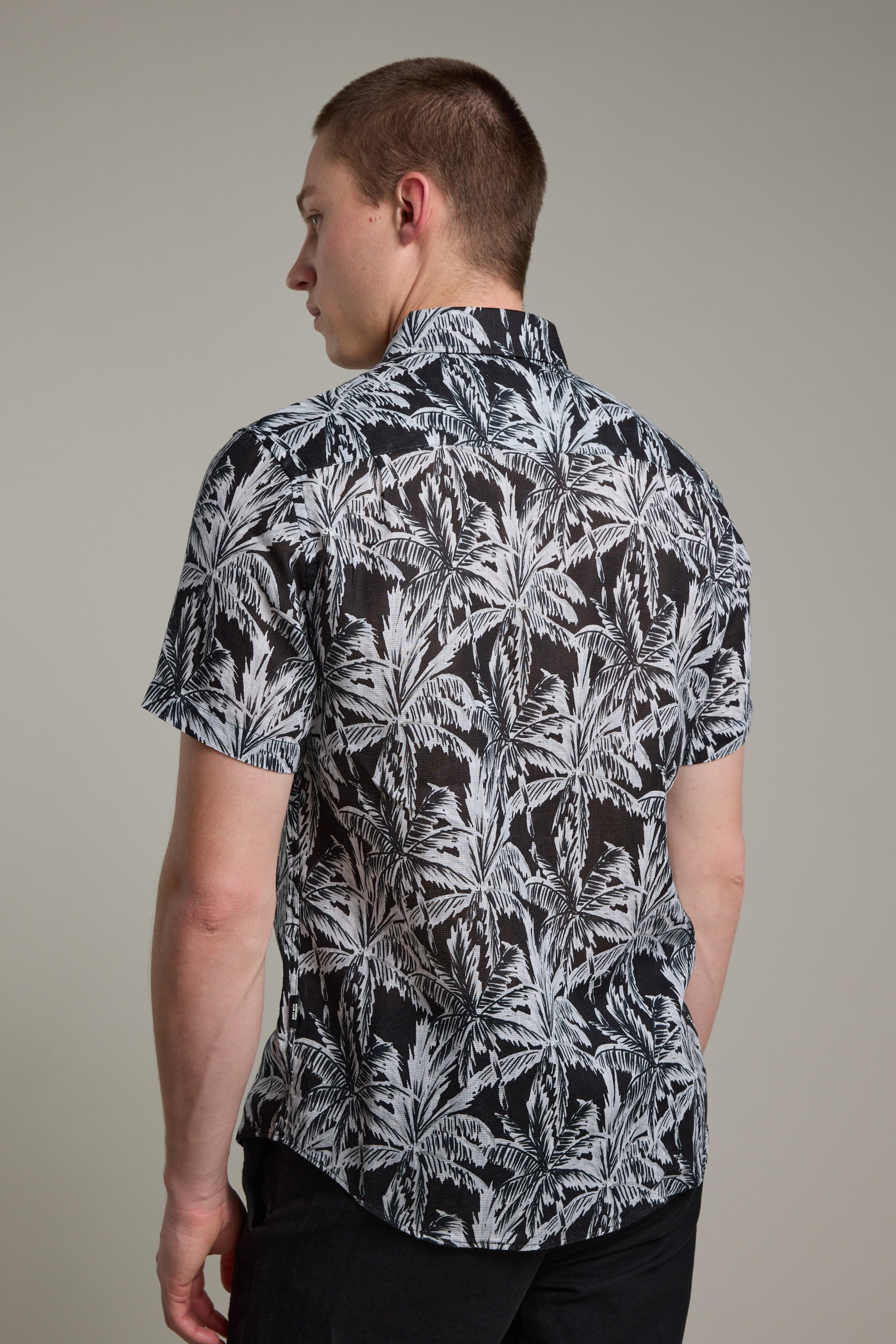 A man faces away, wearing Barkers' Madison Print Shirt—a short-sleeve black Hawaiian shirt with a white palm leaf pattern in soft cotton-linen blend—paired with dark pants against a plain light gray background.