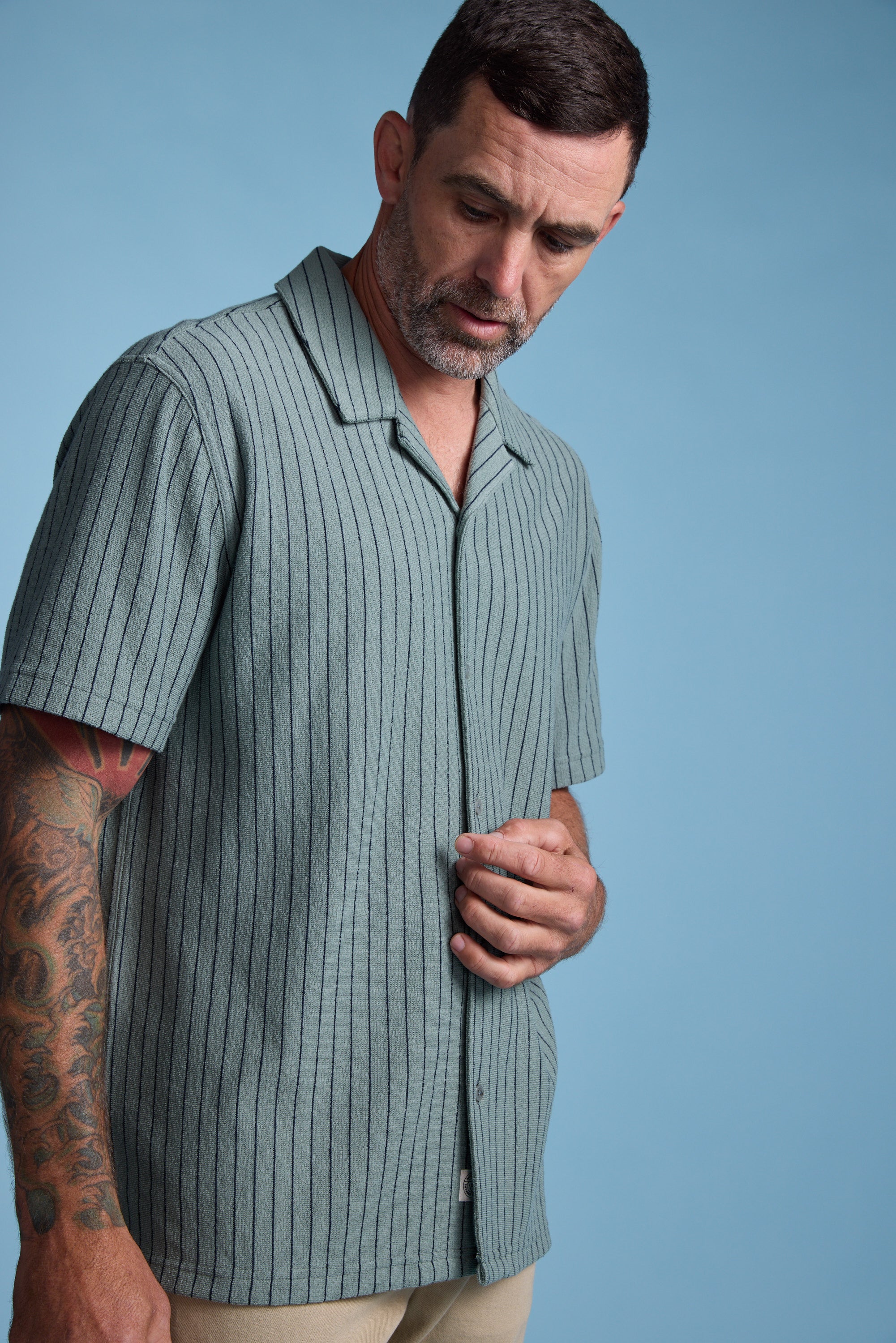 A man with short dark hair, a beard, and tattoos on his left arm stands against a plain blue background, looking down while buttoning the Barkers Diego Stripe Cuban Shirt—a light green, boxy-fit shirt with vertical stripes.