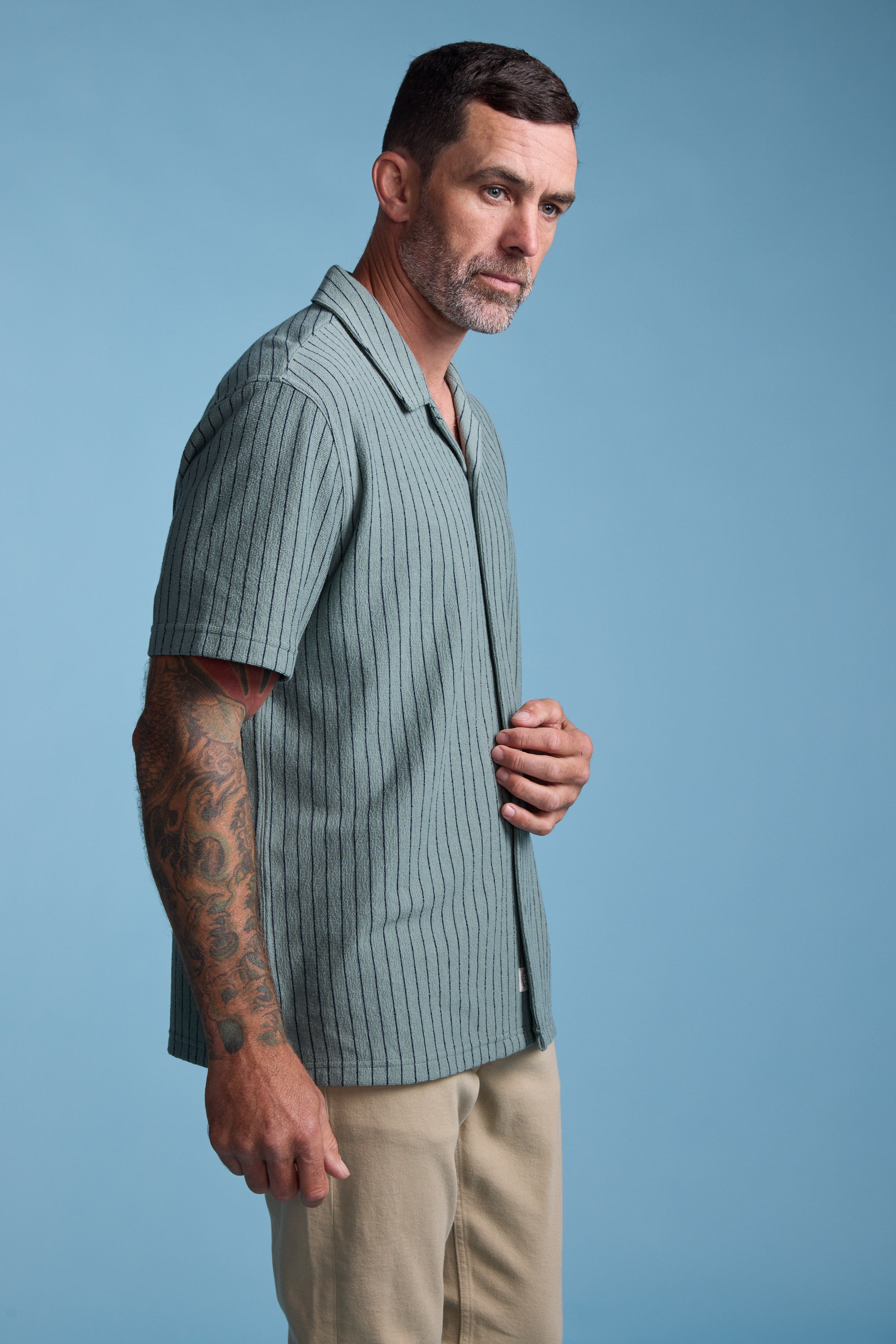 A man with short dark hair and a beard wears the Barkers Diego Stripe Cuban Shirt—a green, boxy-fit style—along with beige pants. Against a blue background, he shows off a tattoo sleeve on his left arm and gazes thoughtfully to the side.