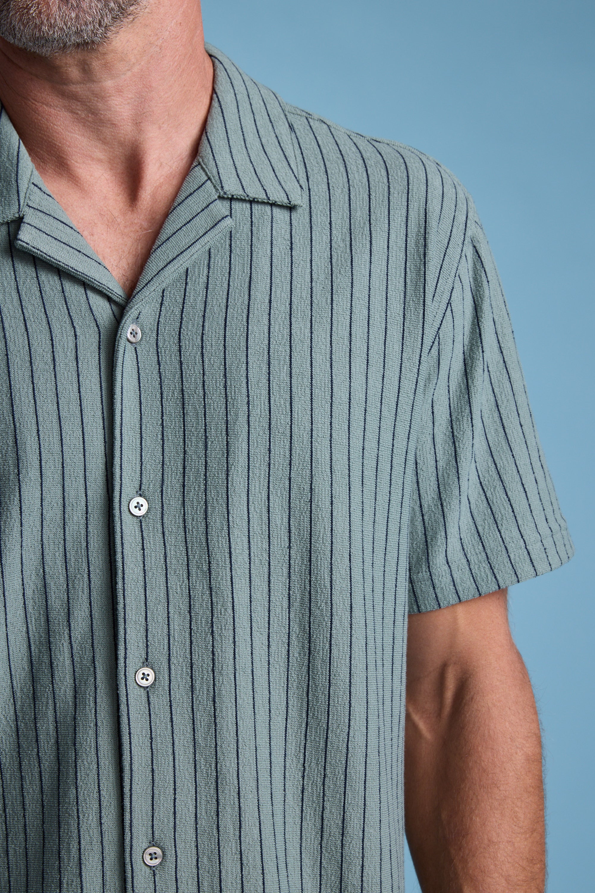 A close-up shows a man in the Barkers Diego Stripe Cuban Shirt—a sage green terry toweling shirt with vertical dark stripes, white buttons, and a boxy fit. Only his lower face, neck, and upper torso are visible against a light blue background.