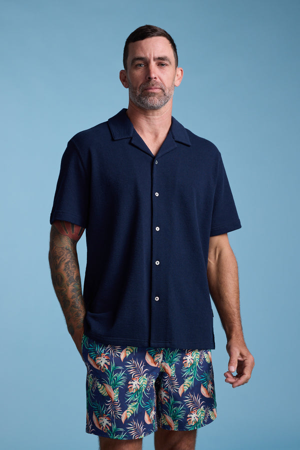 A man with short dark hair, a beard, and tattoos on his left arm stands against a blue background wearing the Barkers Diego Cuban Shirt—a navy short-sleeve shirt—paired with colorful tropical-patterned shorts.
