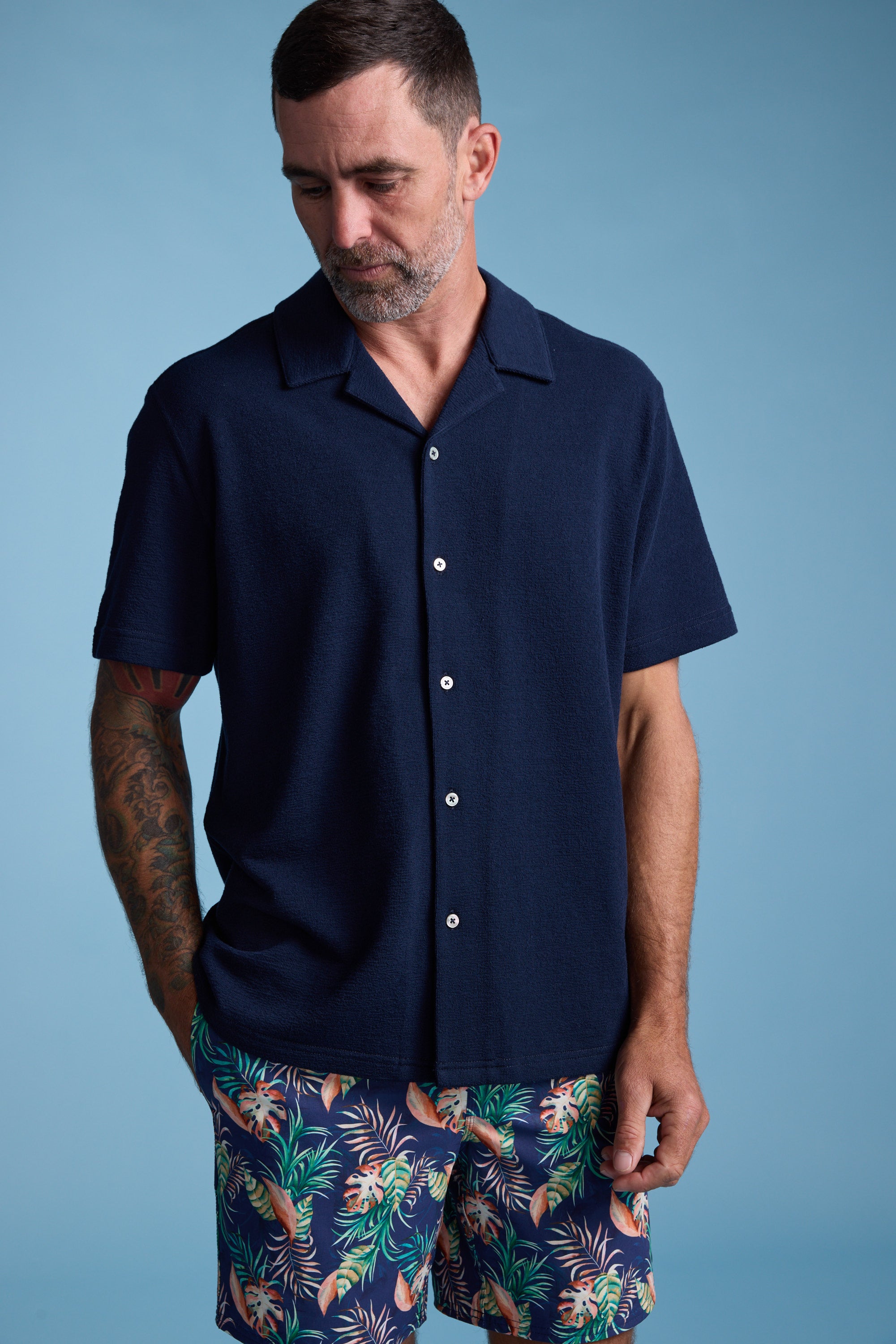 A man with short dark hair and a beard wears the Barkers Diego Cuban Shirt, styled with colorful floral shorts, standing against a plain blue background with one hand in his pocket while looking down.