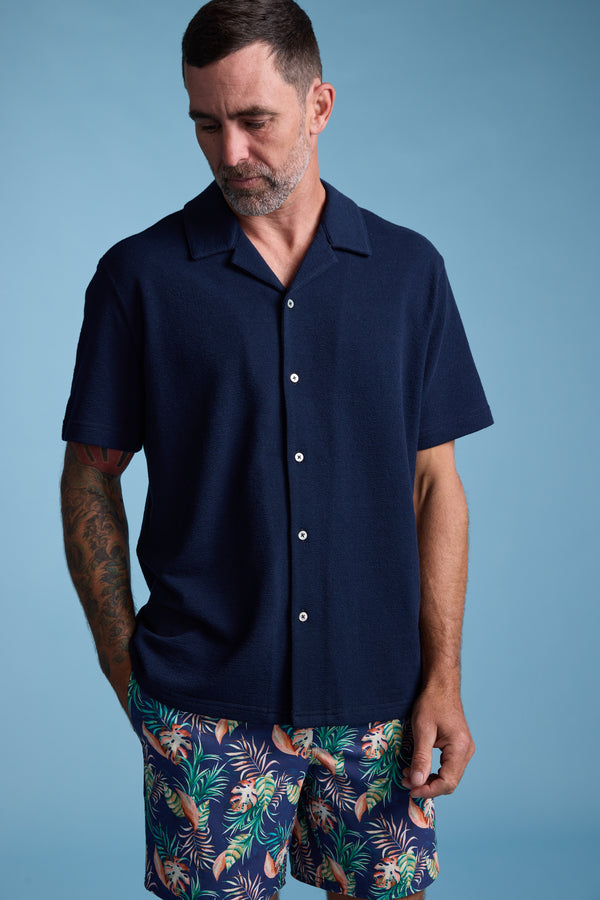 A man with short dark hair and a beard wears the Barkers Diego Cuban Shirt, styled with colorful floral shorts, standing against a plain blue background with one hand in his pocket while looking down.