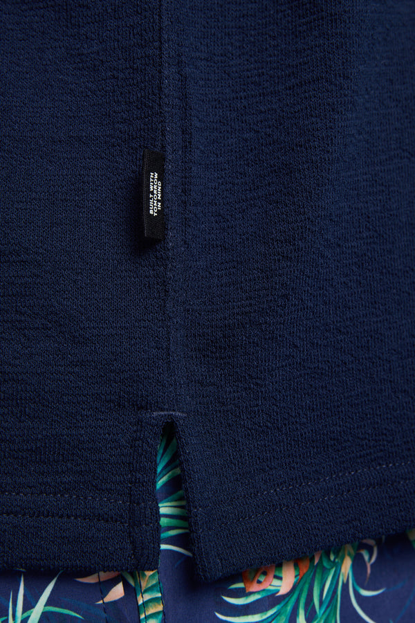 A close-up of the Barkers Diego Cuban Shirt in navy textured terry toweling with a boxy fit and small black tag near the hem, paired with blue shorts featuring a green and orange tropical leaf pattern.