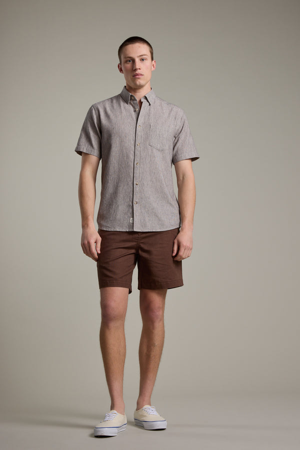 A man stands against a plain background, wearing a short-sleeve, button-up beige Barkers Woodley Texture Shirt, brown shorts, and white sneakers. He faces forward with a neutral expression and his arms at his sides.