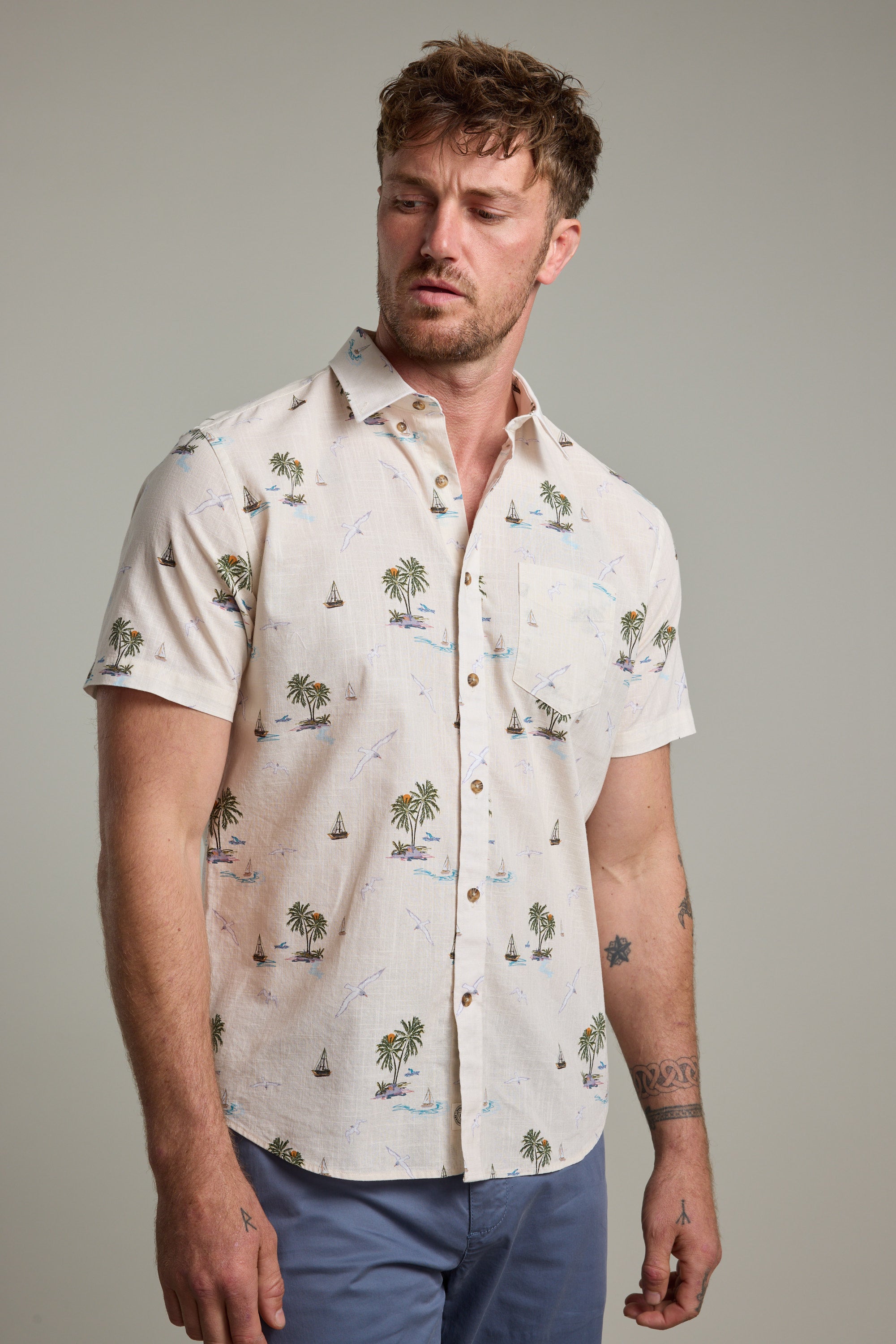 A man with tousled hair models the Barkers Island Print Shirt, a light-colored short sleeve shirt with palm trees and sailboats, standing against a plain background and gazing left for an effortlessly relaxed summer look.