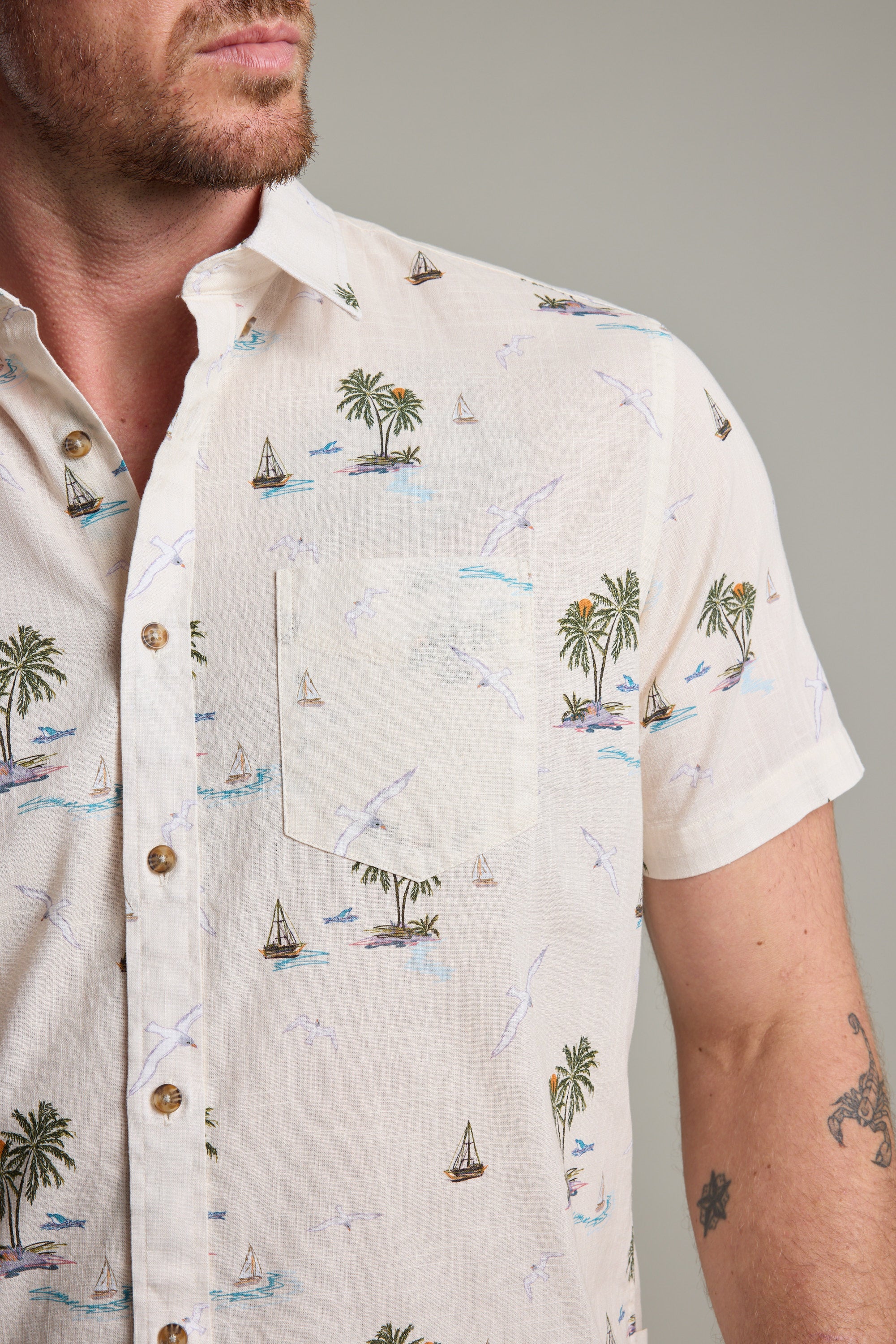 A man wears the Barkers Island Print Shirt, a short sleeve light-colored top with palm trees, sailboats, and birds—a perfect summer style. Only his lower face, neck, and upper torso are shown.