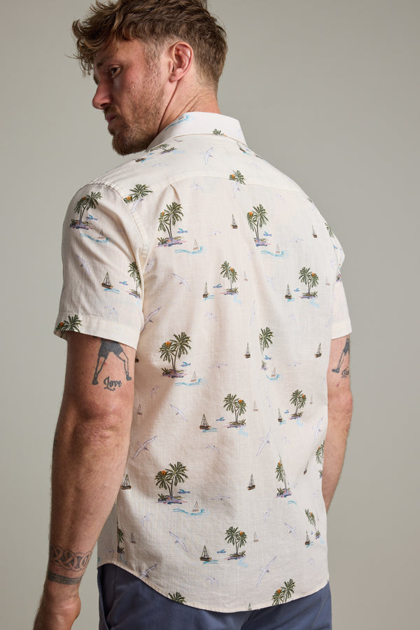 A man with short, curly hair and arm tattoos models the Barkers Island Print Shirt, featuring palm trees and sailboats—ideal for summer. He stands slightly turned from the camera, glancing over his shoulder.