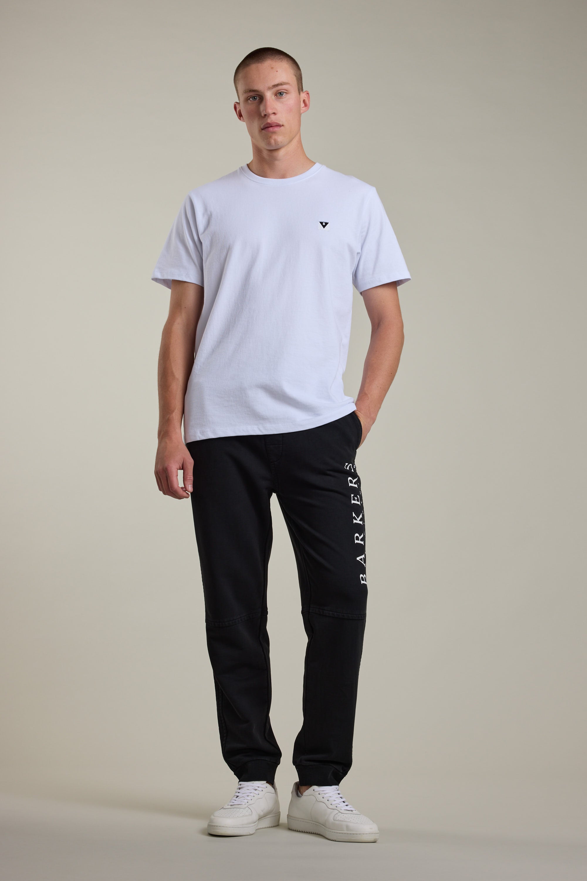 A man with short hair and one hand in his pocket stands against a plain light background, wearing a white t-shirt, Barkers Organic Classic Print Track Pant with BARKERS on the leg, and white sneakers.