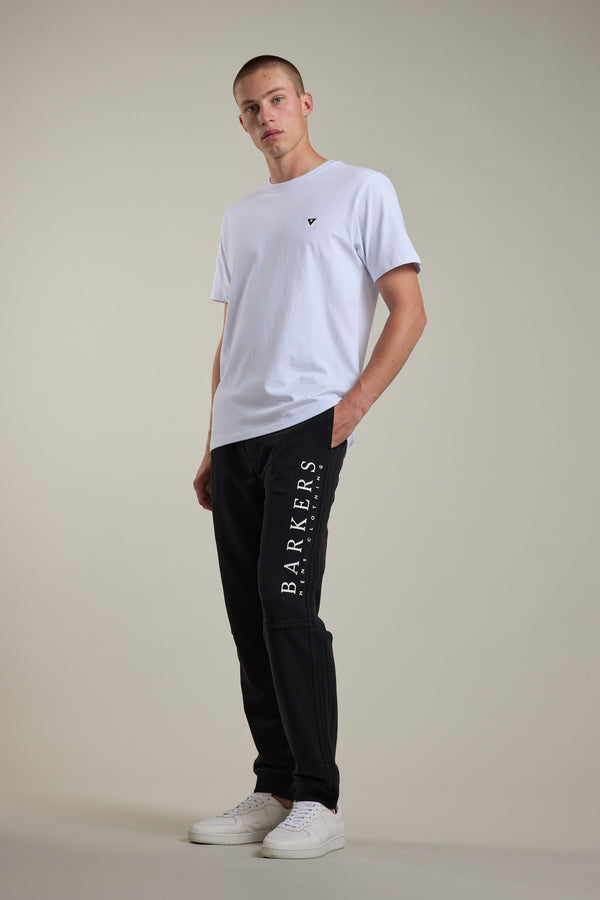 A person poses against a plain background in a white t-shirt, Barkers Organic Classic Print Track Pant with 'BARKERS' along the leg, and white sneakers—one hand in their pocket, evoking classic ‘90s trackies style.