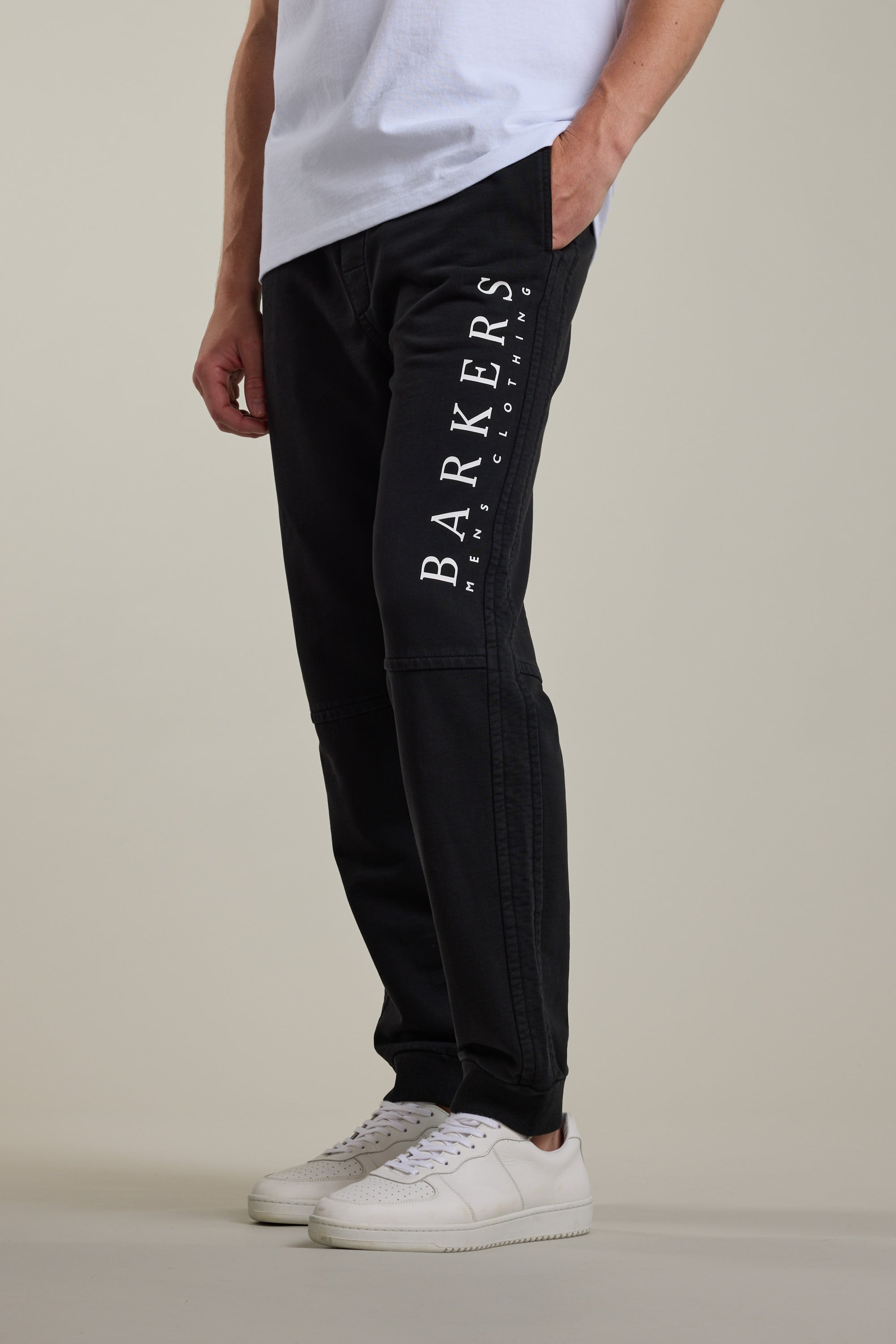 A person wears Barkers Organic Classic Print Track Pants with BARKERS MENS CLOTHING in white on the leg, paired with a white T-shirt and sneakers, standing against a plain background.