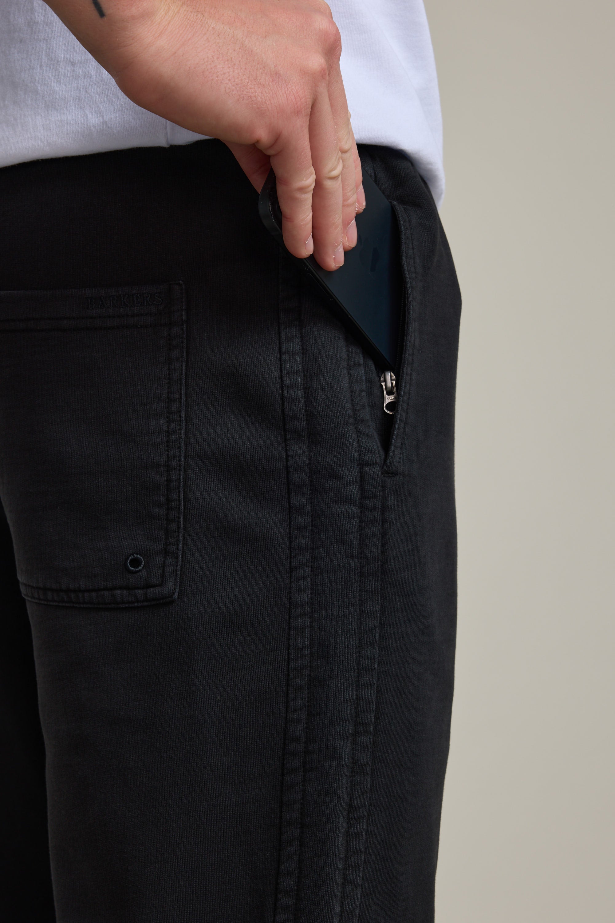 Someone places a black smartphone into the zippered pocket of their Barkers Organic Classic Print Track Pant. The person's upper body and part of a white shirt are also visible.