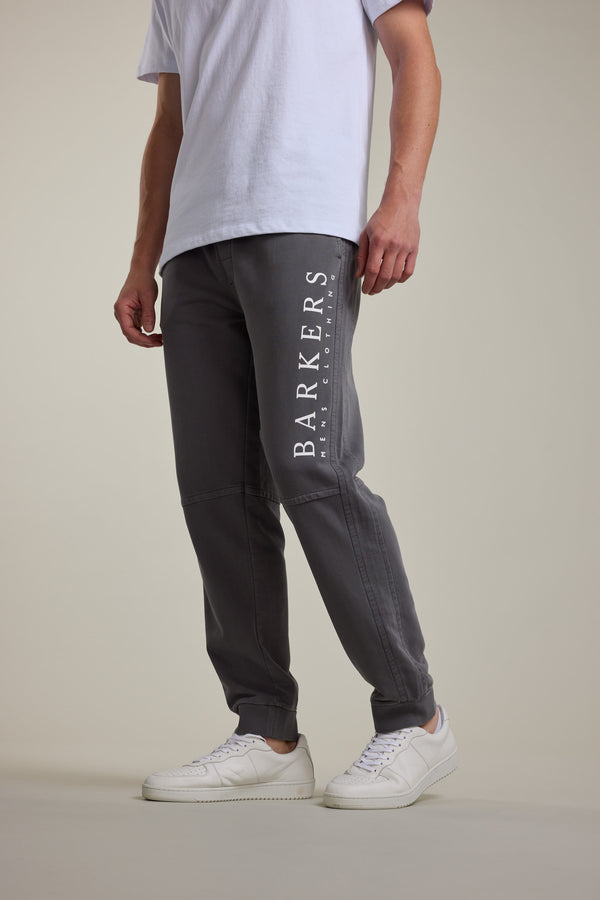 A person wearing a white T-shirt, Barkers Organic Classic Print Track Pant with BARKERS branding on the left leg, and white sneakers stands against a plain light background.
