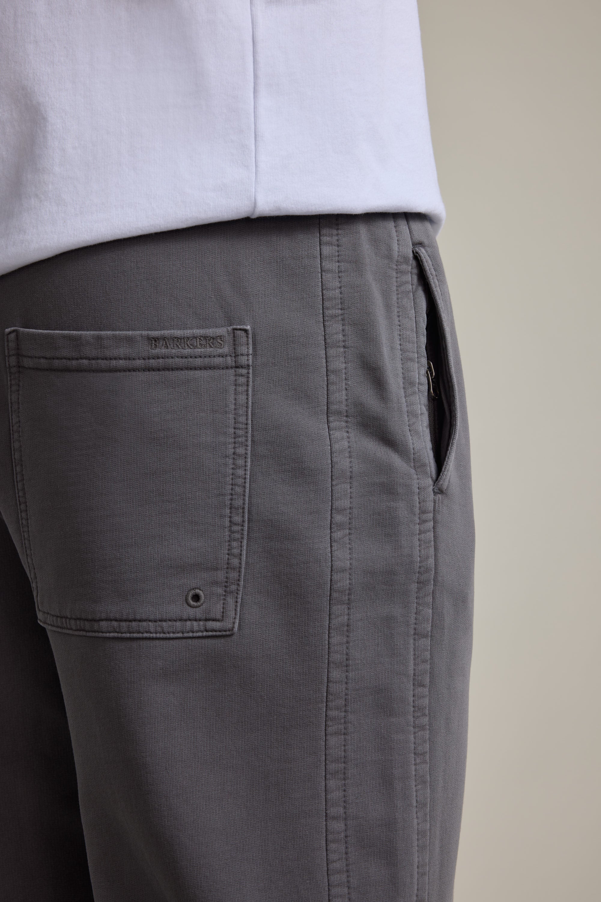 Close-up of a person wearing Barkers Organic Classic Print Track Pant in gray, featuring a side pocket, visible stitching, and the Barkers logo stitched above the back pocket. Paired with a white shirt against a neutral background.