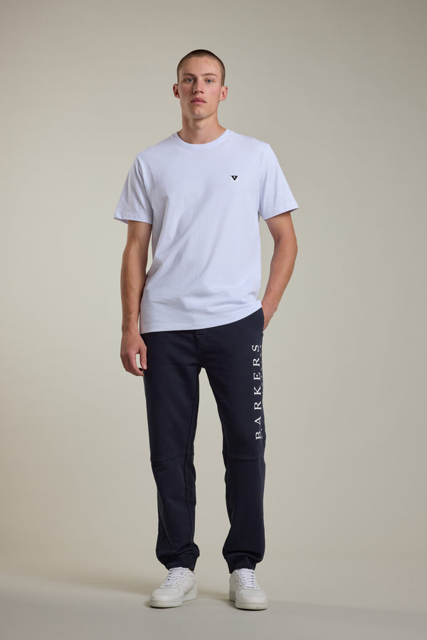 A young man stands against a plain light background, wearing a white t-shirt, white sneakers, and Barkers Organic Classic Print Track Pant with BARKERS branding on the left leg. One hand is in his pocket as he faces the camera neutrally.