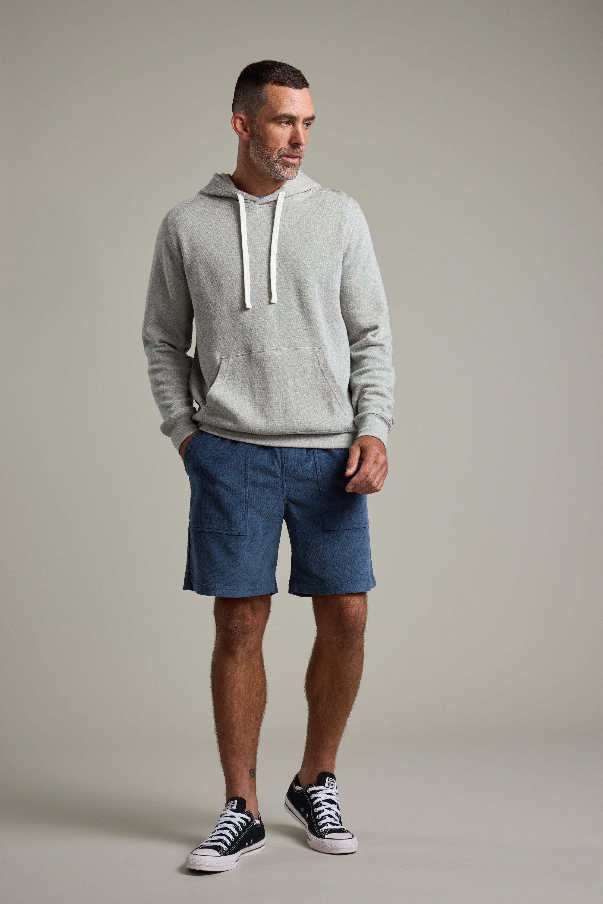 A man with short hair and a beard stands against a plain background wearing the Barkers Inlet Hoody in light gray, blue shorts, and black low-top sneakers with white soles, his hands tucked in the hoody's pockets.