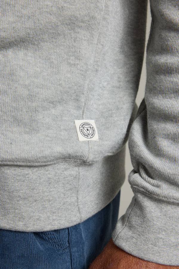 A close-up shows part of a person’s arm and hand wearing the Barkers Inlet Hoody in light grey organic cotton, featuring ribbed sleeves and a small white tag near the hem, paired with blue pants.