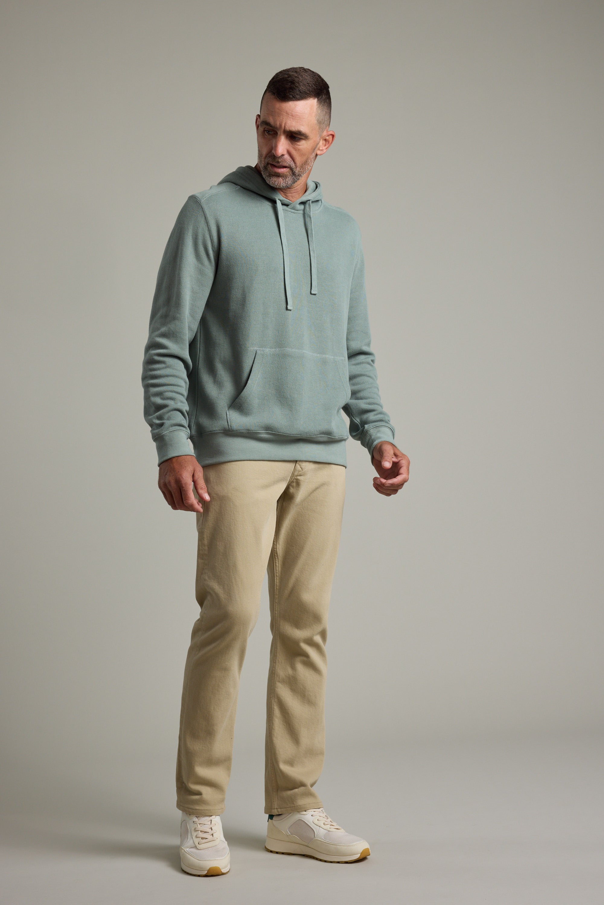 A man wears the Barkers Inlet Hoody in light blue, paired with beige pants and white sneakers, standing against a plain light gray background and gazing slightly to his left with a neutral expression.