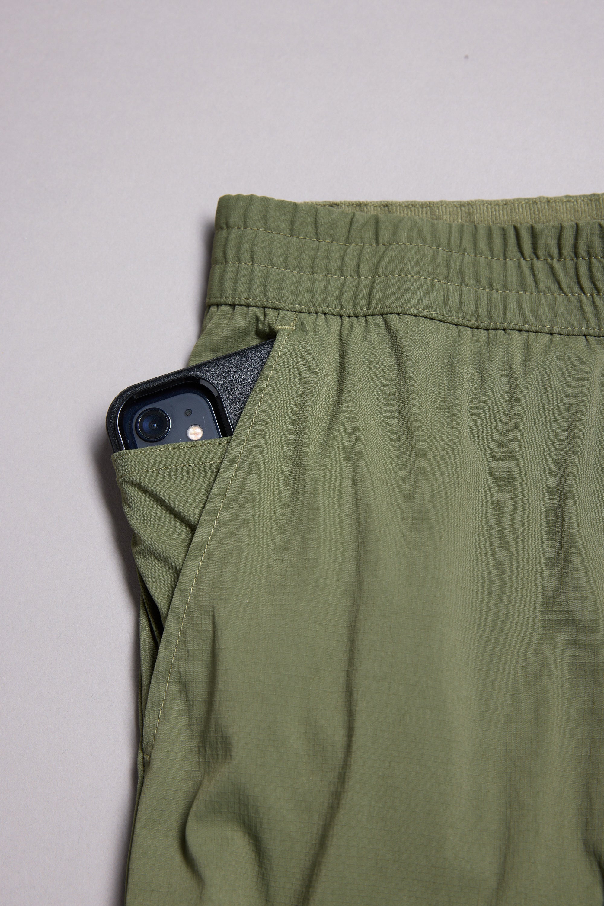 A black smartphone peeks from the side pocket of olive green WearEver Transit Pants by WearEver, featuring an elastic waist, shown against a neutral background.