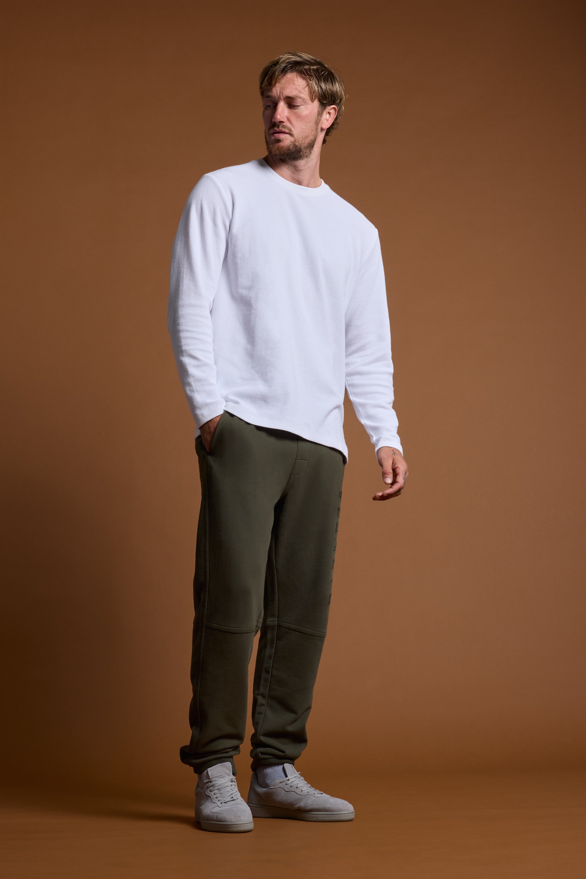 A man stands against a plain brown background, wearing Barkers Heritage Track Pant in olive green french terry organic cotton with white long-sleeve shirt and light gray sneakers, showcasing the Barkers logo detail as he looks to the side with one hand in his pocket.