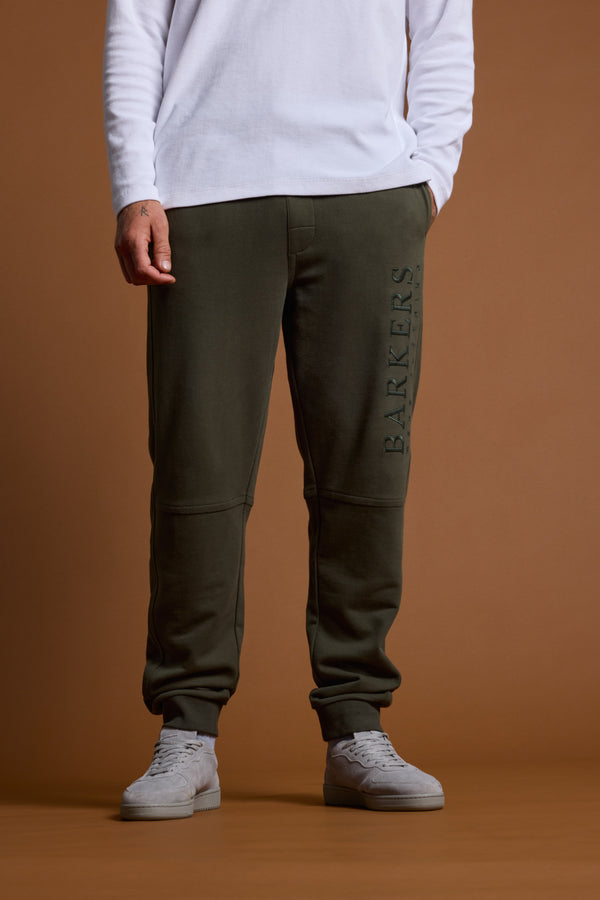 A person stands against a brown background, wearing a white long-sleeve shirt, Barkers Heritage Track Pant in dark green with the Barkers logo on the left leg, and light gray sneakers.