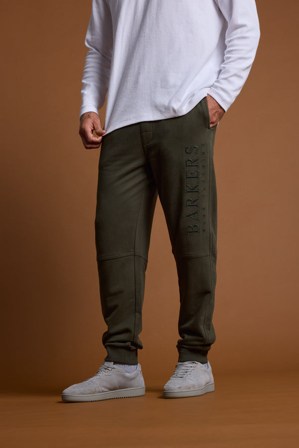A person in a white long-sleeve shirt, Barkers Heritage Track Pant in dark green with the Barkers logo, and light gray sneakers stands against a plain brown background.