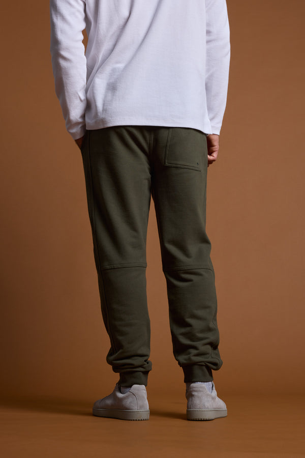 A person stands facing away, wearing a white long-sleeve shirt, Barkers Heritage Track Pant joggers by Barkers with a back pocket, and light gray sneakers, set against a plain brown background.