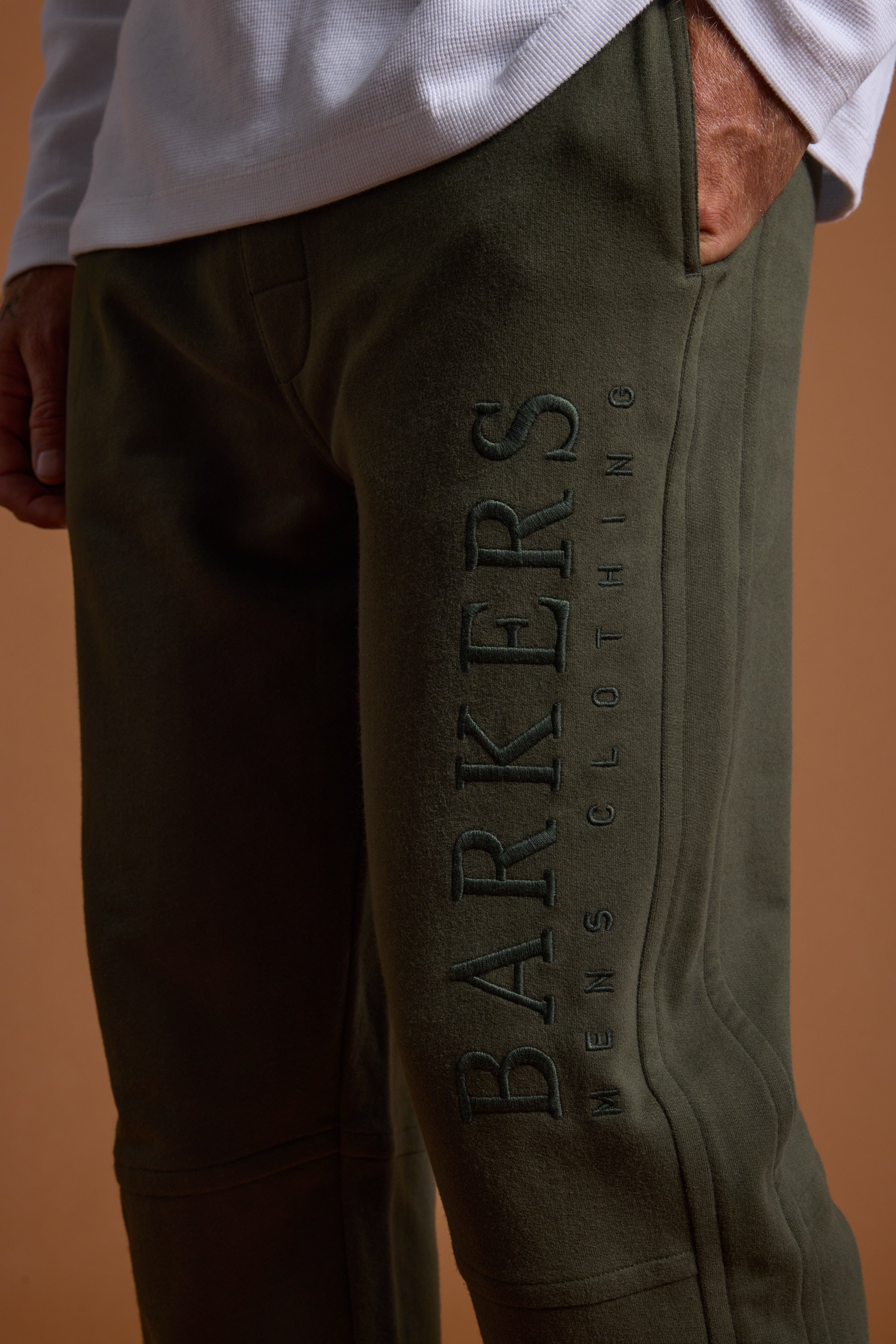 A person wears Barkers Heritage Track Pant in olive green organic cotton, with one hand in the pocket and “BARKERS” heritage logo embroidered vertically on the side. The warm brownish background highlights the comfy French terry fabric.
