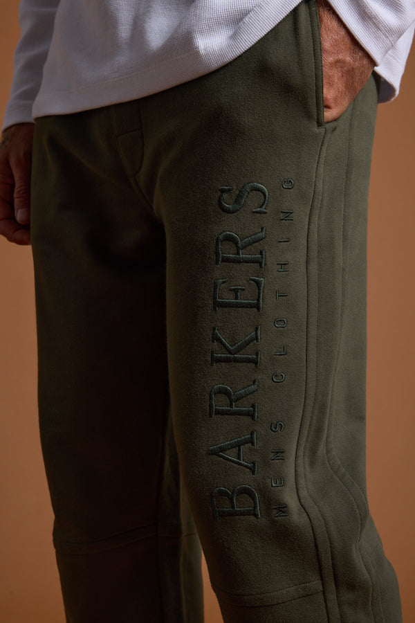 A person wears Barkers Heritage Track Pant in olive green organic cotton, with one hand in the pocket and “BARKERS” heritage logo embroidered vertically on the side. The warm brownish background highlights the comfy French terry fabric.