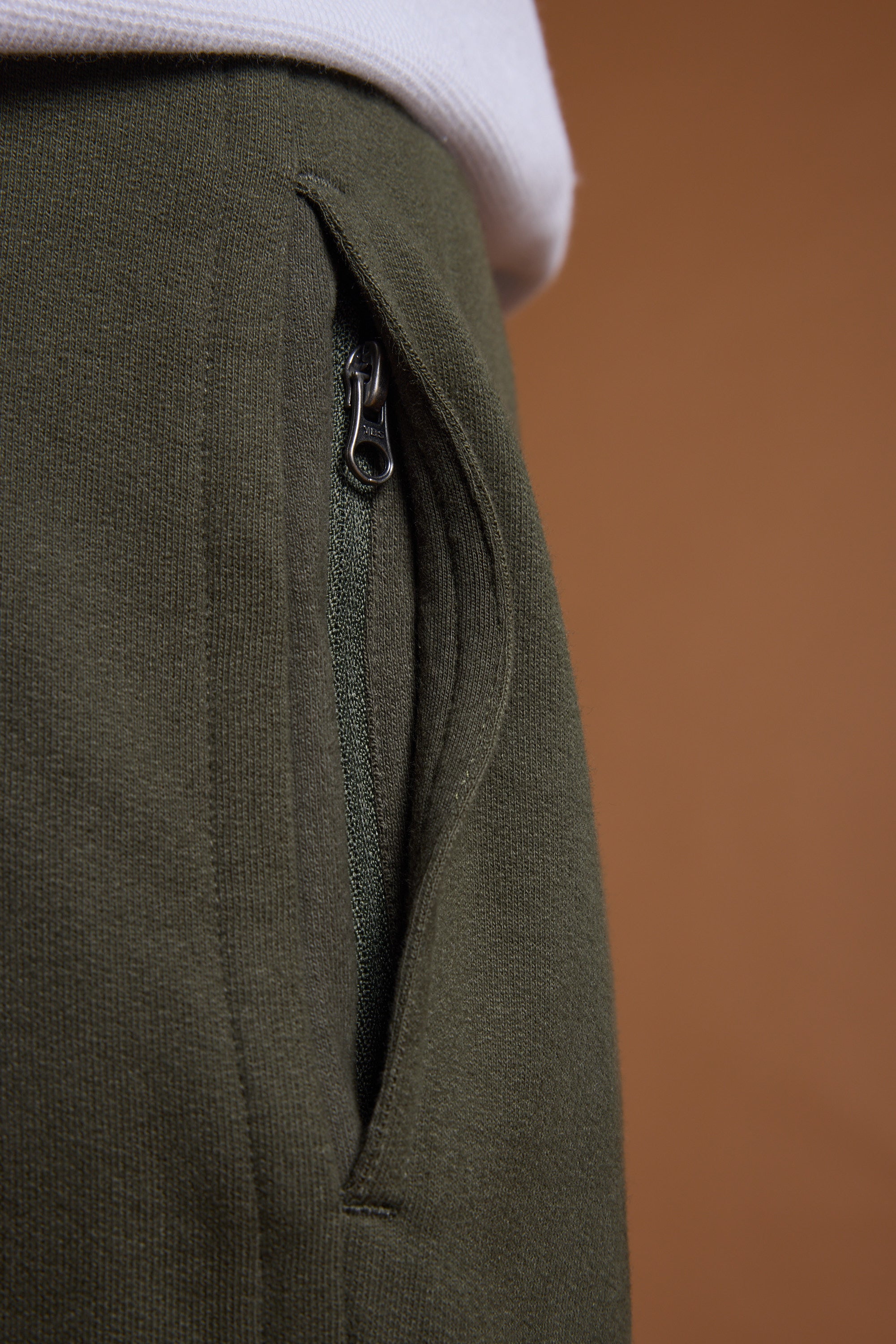 Close-up of a person wearing Barkers Heritage Track Pant in olive green organic cotton with a partially open zippered pocket and the Barkers heritage logo, paired with a white shirt against a solid brown background.