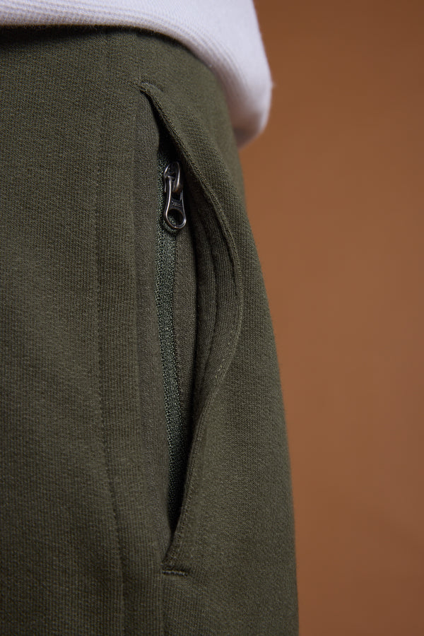 Close-up of a person wearing Barkers Heritage Track Pant in olive green organic cotton with a partially open zippered pocket and the Barkers heritage logo, paired with a white shirt against a solid brown background.