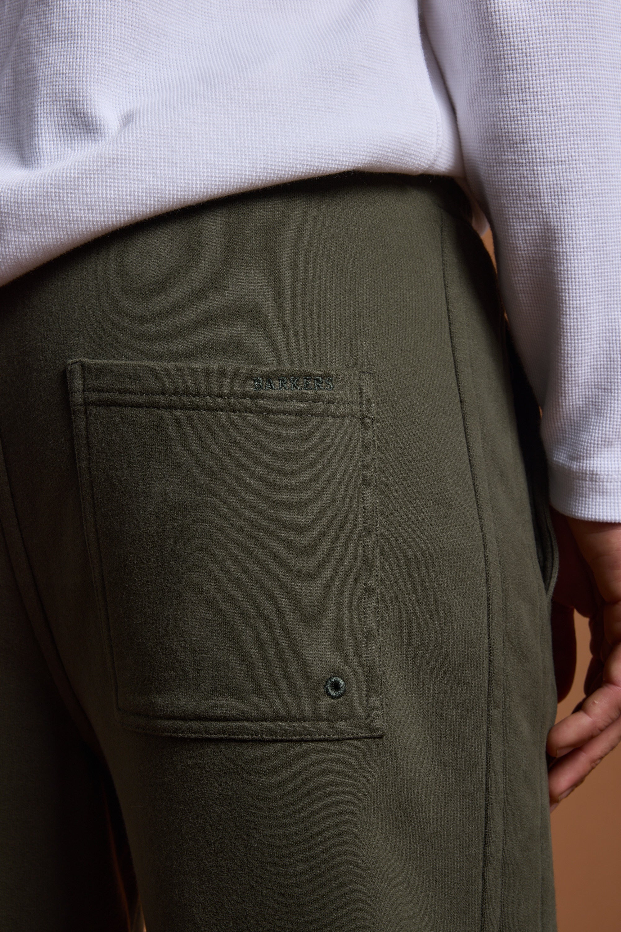 Close-up of a person wearing Barkers Heritage Track Pant by Barkers in olive green organic cotton, featuring a back pocket with metal grommet, paired with a white textured long-sleeve shirt. Only the waist and upper leg are shown.