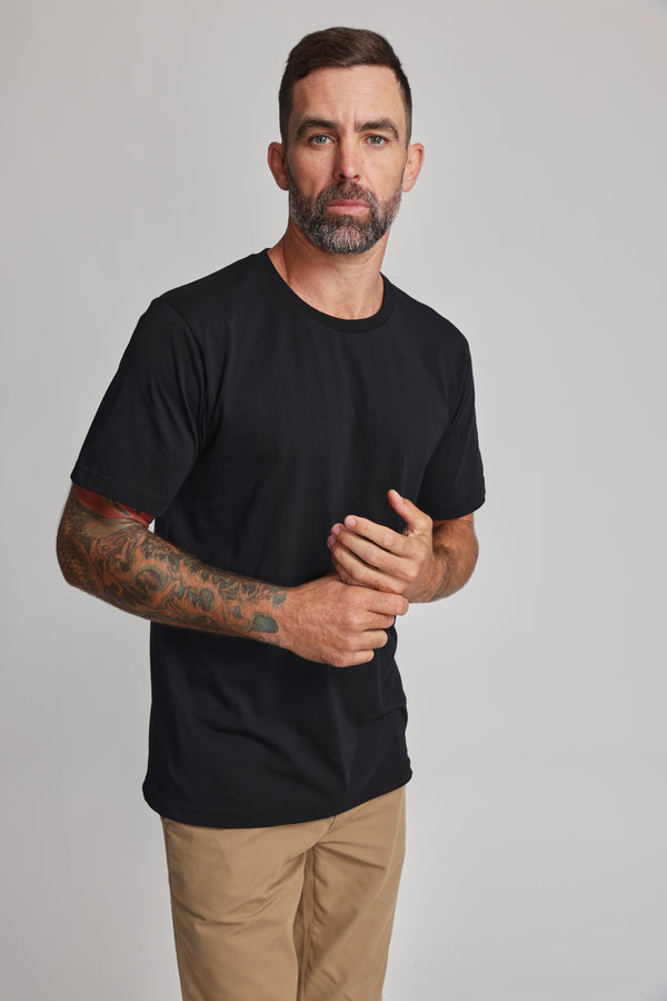 A man with short dark hair, a beard, and visible left forearm tattoos wears a black Barkers Organic Crew Tee and tan pants, standing against a plain light gray background.