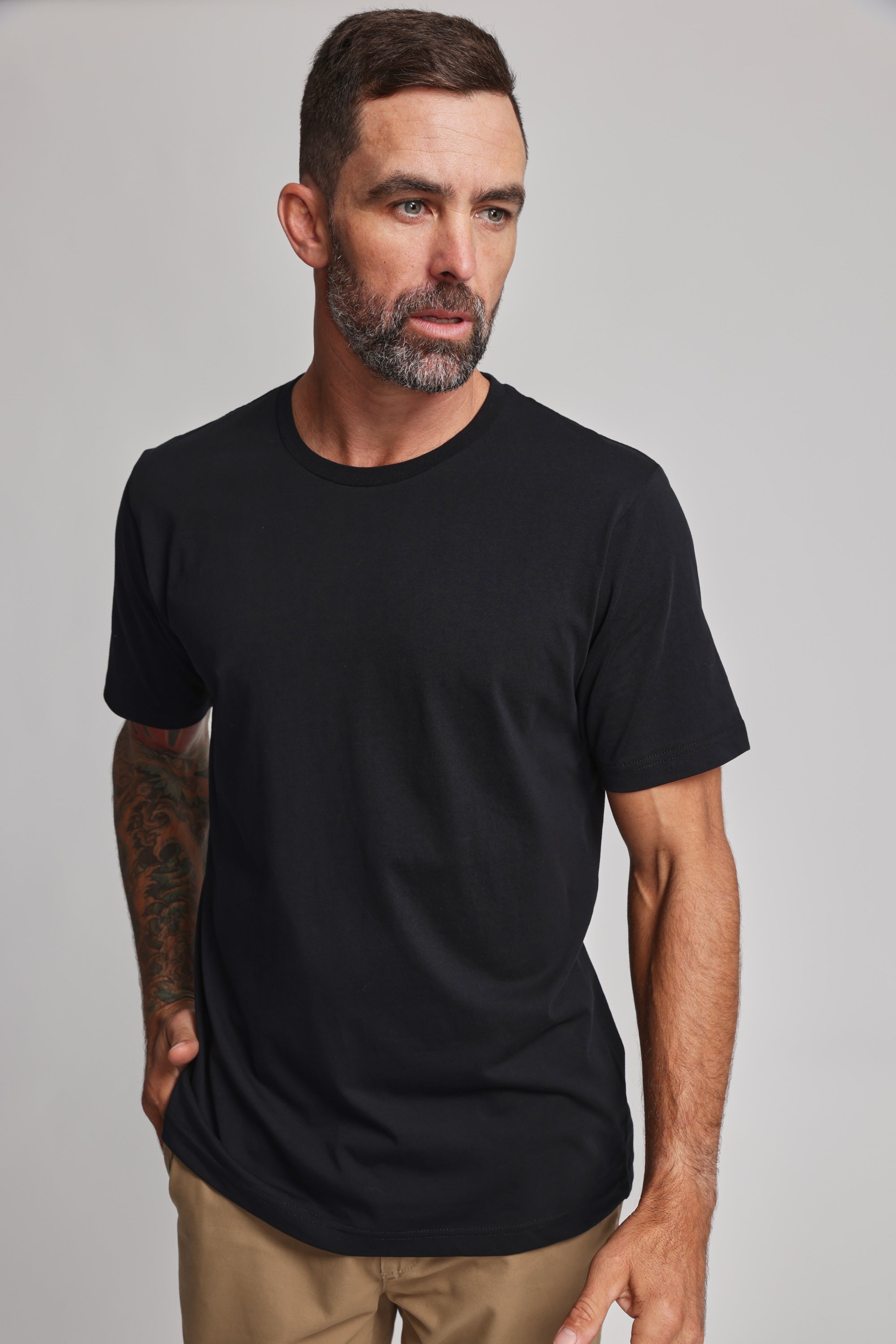 A man with short dark hair and a beard wears the Barkers Organic Crew Tee, a plain black 100% organic cotton mid-weight top, paired with khaki pants as he stands against a light gray background, looking thoughtfully to his right.