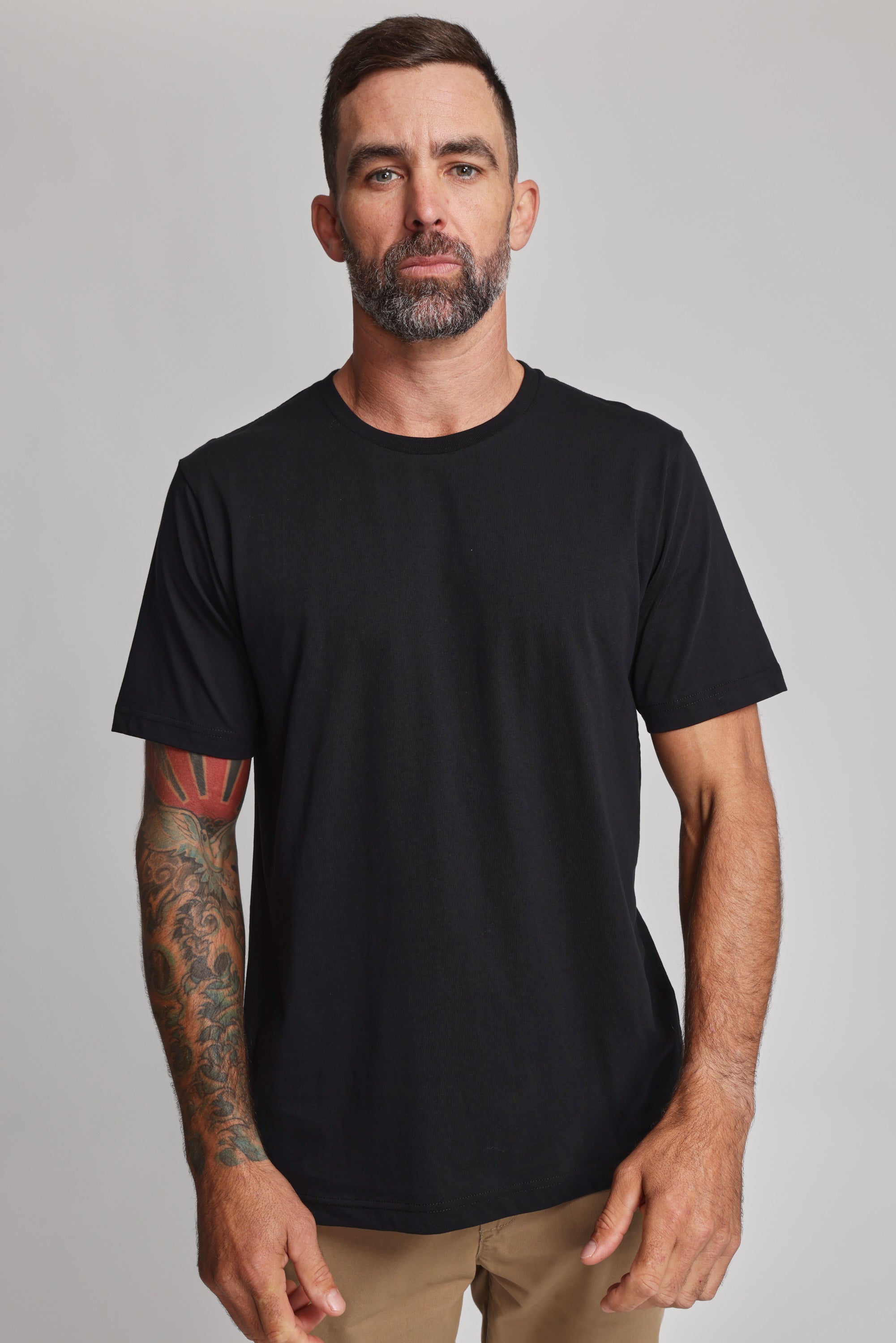 A man with short dark hair, a beard, and tattoos on his right arm wears the Barkers Organic Crew Tee—plain black and made from 100% organic cotton—with tan pants, standing against a light gray background and looking at the camera neutrally.