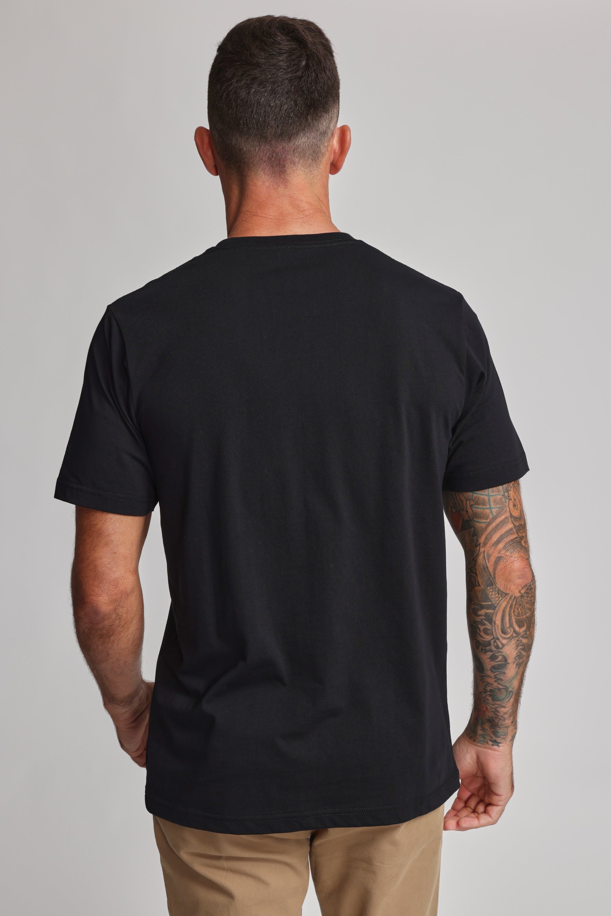 A man with short dark hair and a tattooed right arm stands with his back to the camera, wearing Barkers' Organic Crew Tee in black—crafted from 100% organic cotton—and khaki pants, set against a plain light background.