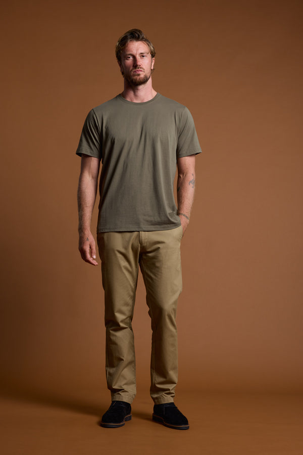 A man stands against a plain brown background, wearing the Barkers Organic Crew Tee in khaki green—crafted from 100% organic cotton—paired with tan pants and black shoes. He has short hair, a neutral expression, and his hands rest at his sides.