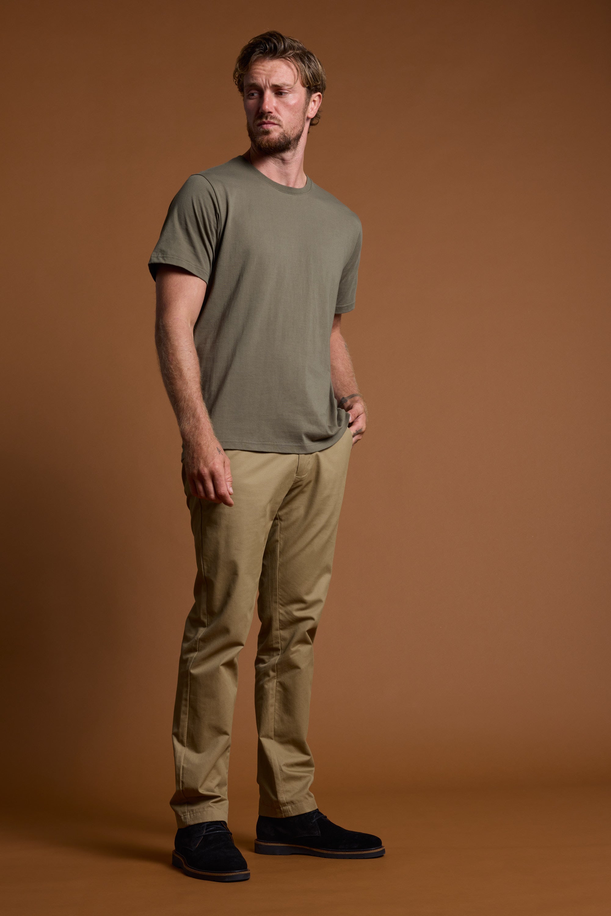 A man with light brown hair and a beard stands against a brown background, wearing Barkers’ mid-weight olive green Organic Crew Tee made from 100% organic cotton, paired with beige pants and black shoes. He looks to the side with a hand in his pocket.