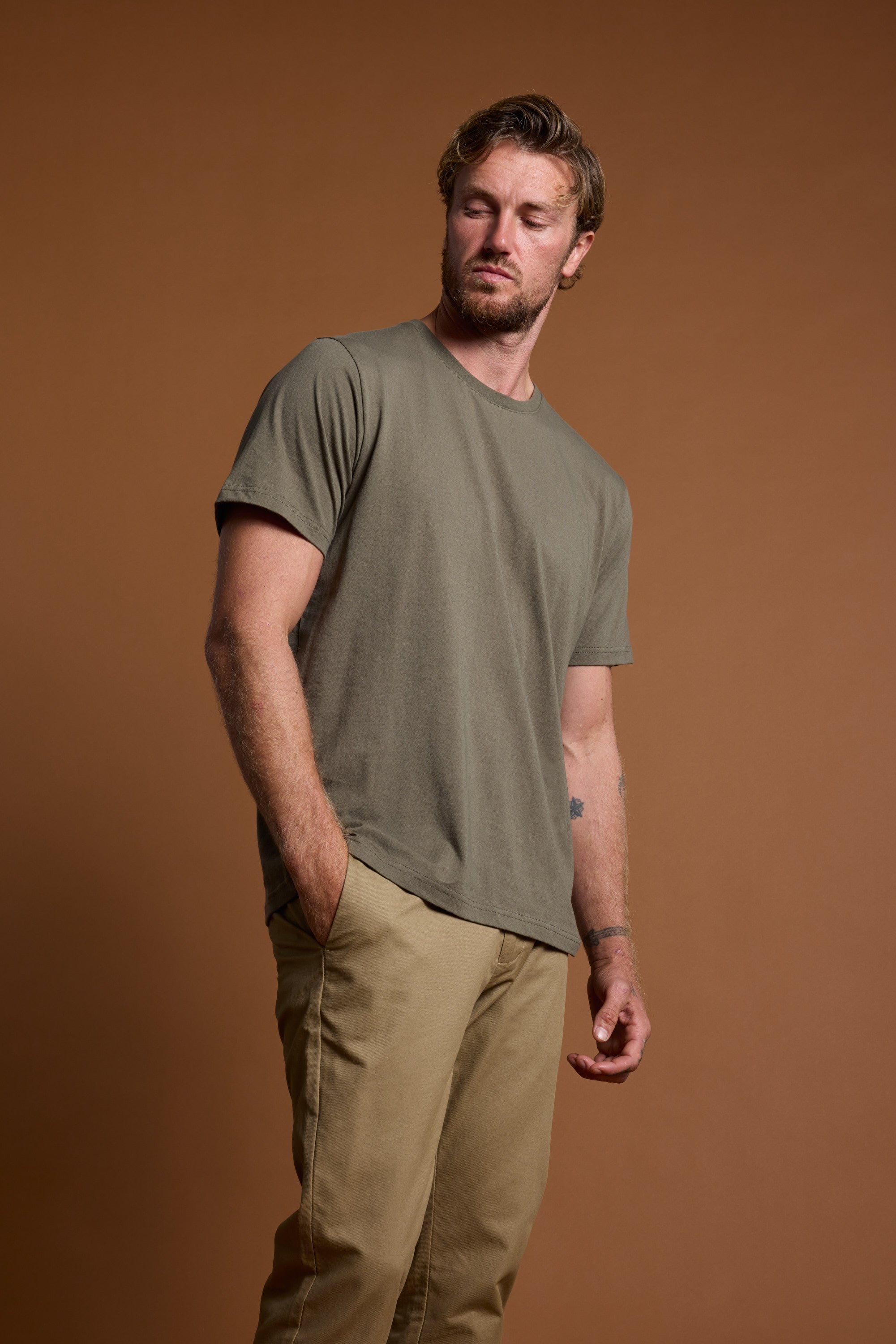 A man with light brown hair and a beard wears the Barkers Organic Crew Tee, made from 100% organic cotton, and beige pants. He stands against a plain brown background with one hand in his pocket, looking slightly to the side.
