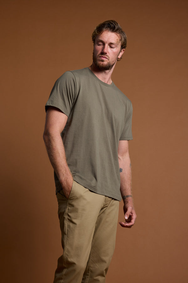 A man with light brown hair and a beard wears the Barkers Organic Crew Tee, made from 100% organic cotton, and beige pants. He stands against a plain brown background with one hand in his pocket, looking slightly to the side.