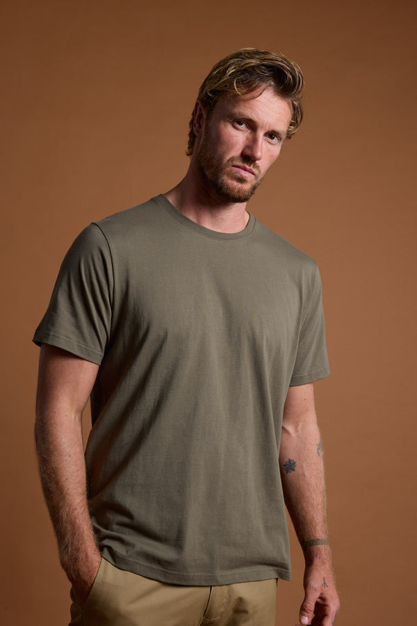 A man with light brown hair and a beard stands against a brown background, wearing Barkers' Olive Green Organic Crew Tee made from 100% organic cotton and beige pants, his hands in his pockets as he looks seriously at the camera.