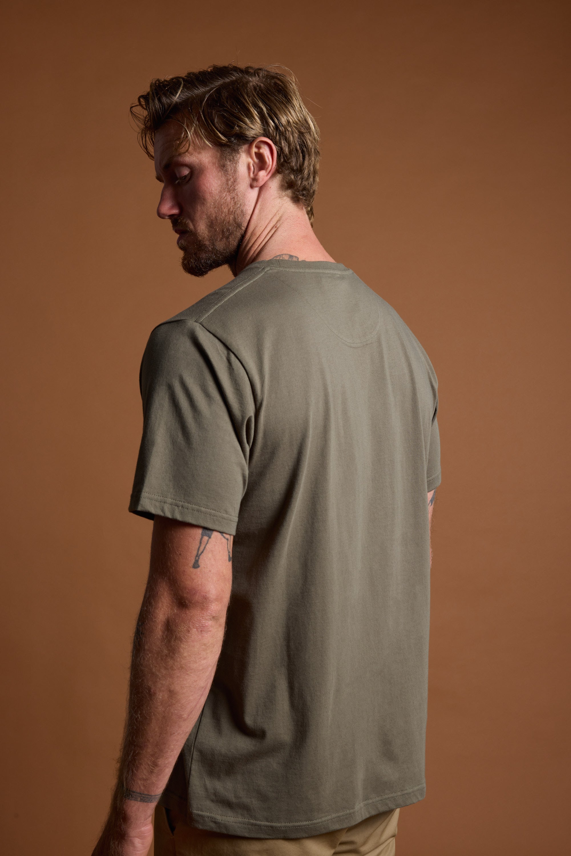 A man with light brown hair and a beard, turned away, wears Barkers' olive green Organic Crew Tee (100% organic cotton) and tan pants. Against a solid brown background, his tattooed left arm is visible.