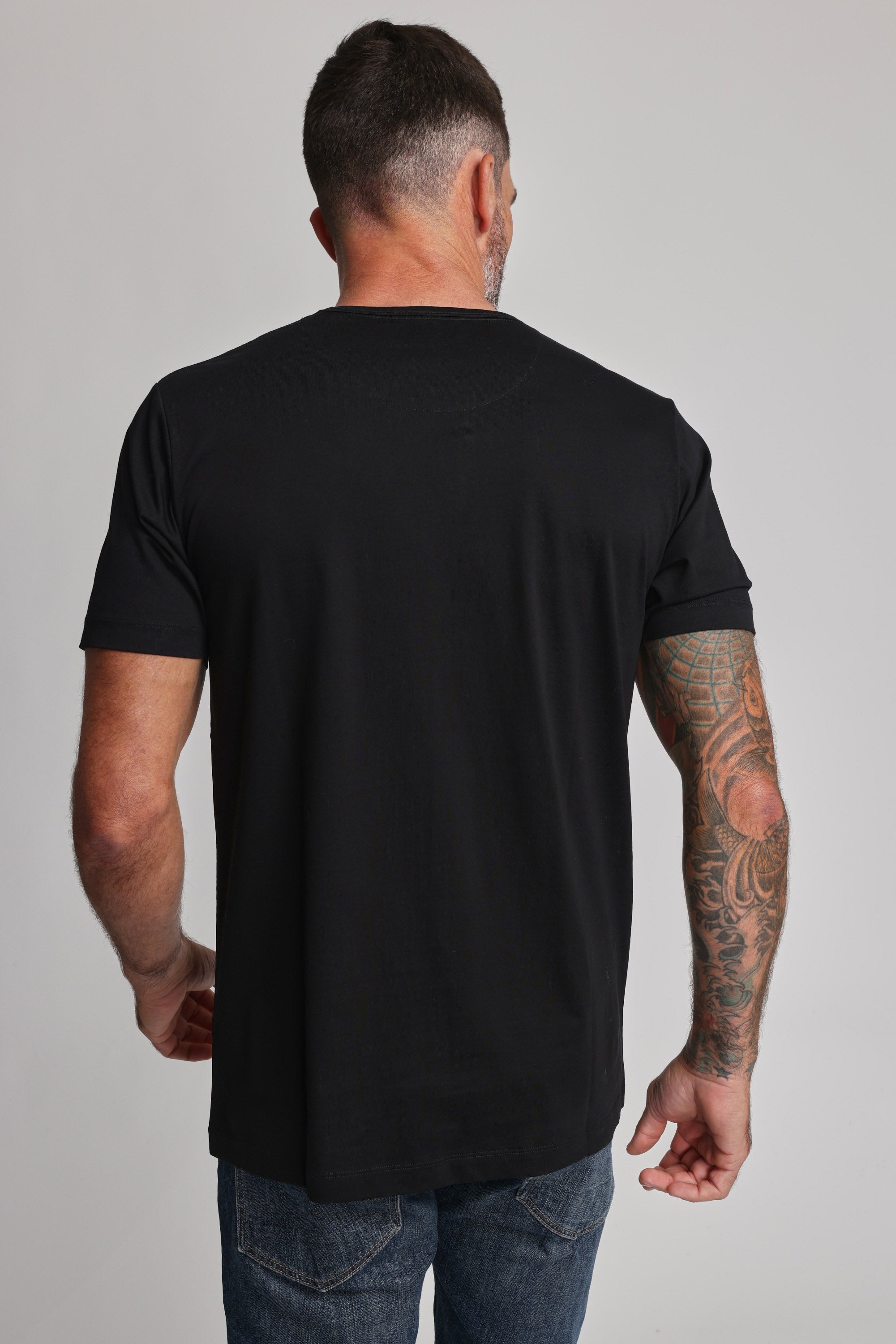 A man with short dark hair and a beard faces away from the camera, wearing a Barkers Linton Supima Tee in black and blue jeans. His right arm displays colorful tattoos against a plain light background.