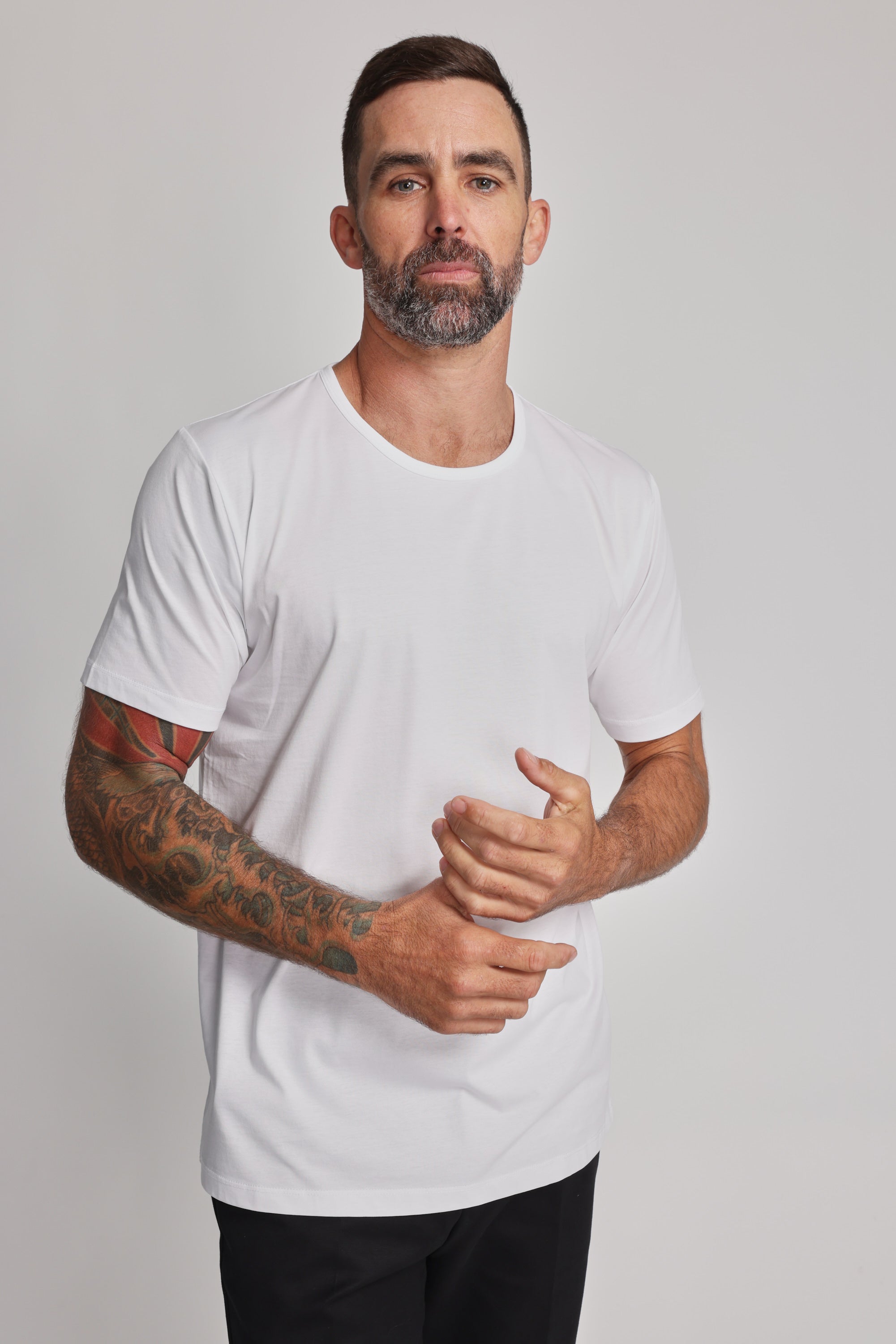 A man with short dark hair, a beard, and right forearm tattoos stands against a plain light background, wearing Barkers’ Linton Supima Tee and black pants—a true wardrobe staple—with his hands clasped together.