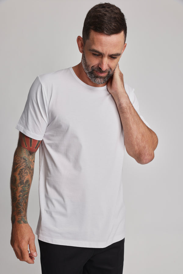 A man with short dark hair and a beard wears the Barkers Linton Supima Tee and black pants. He looks down, touching his neck, showing a colorful tattoo sleeve on his left arm; the background is plain and light-colored.