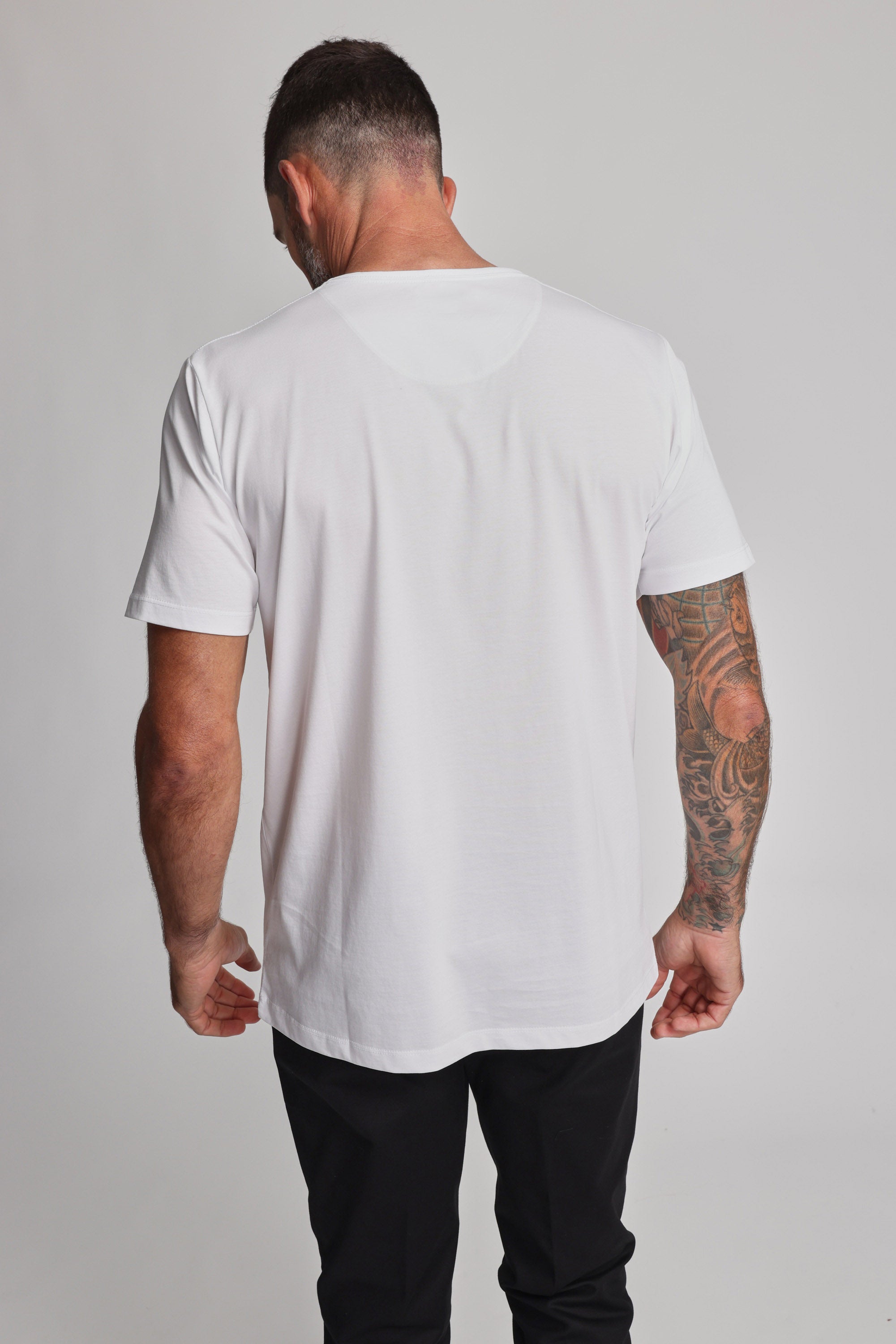 A man with short dark hair and a tattooed right arm stands with his back to the camera, wearing a plain white Barkers Linton Supima Tee and black pants against a light gray background.