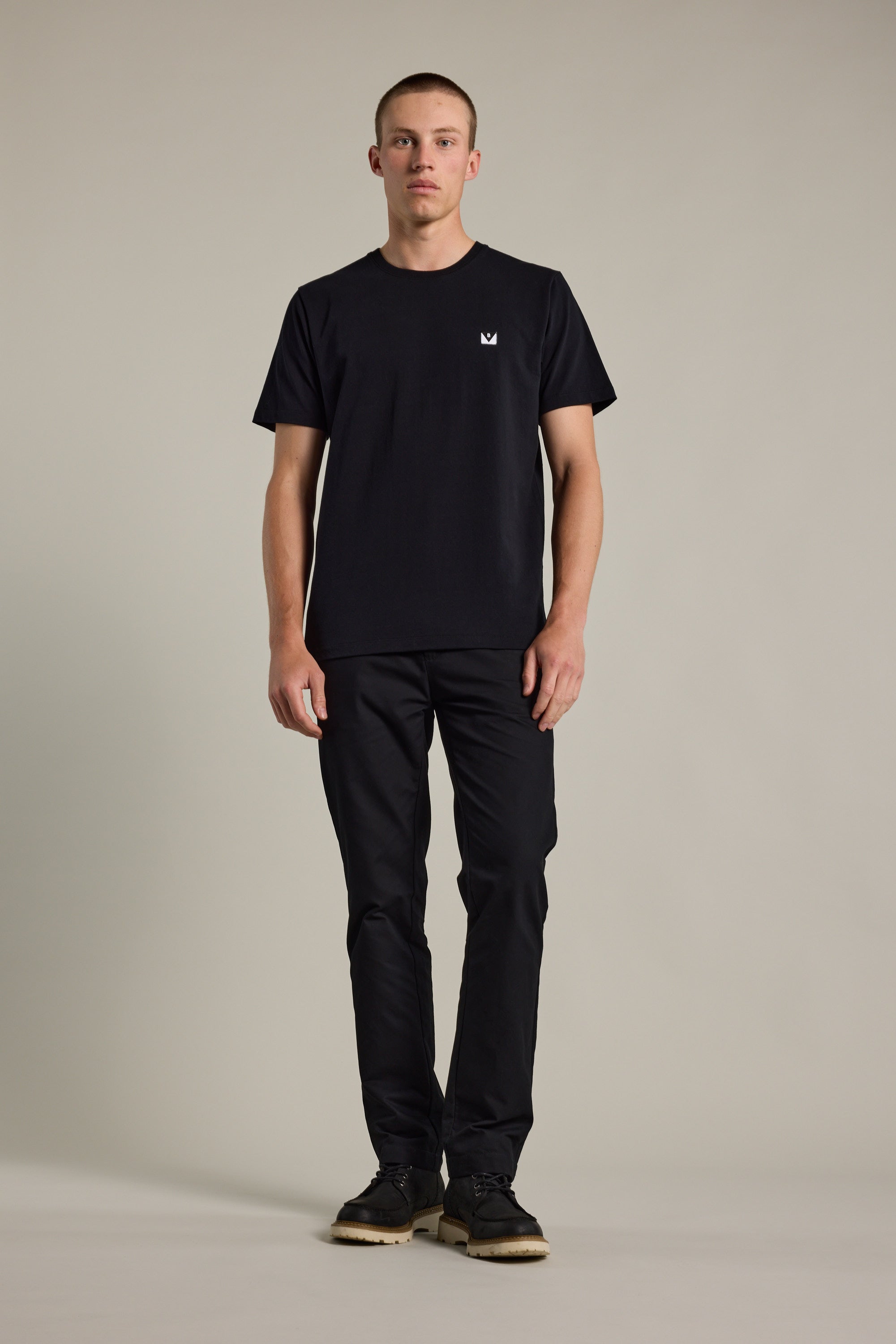 A man stands facing forward against a plain background, wearing the Barkers Newton Washed Tee—a black oversized box fit shirt with a small white logo on the chest—paired with black pants and black shoes.