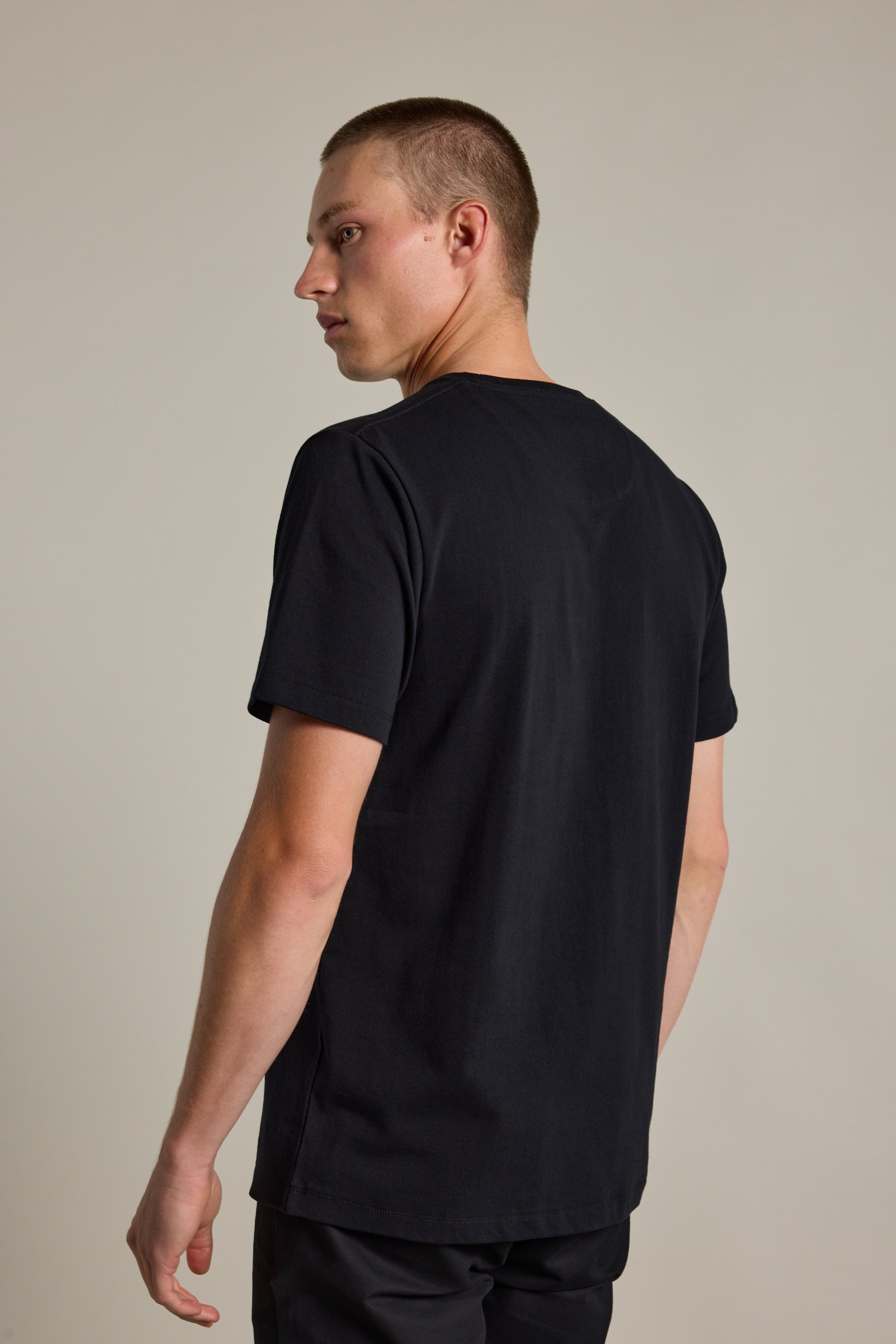 A person with short hair models the Barkers Newton Washed Tee in black, styled oversized with a box fit, standing and looking over their shoulder against a neutral background.