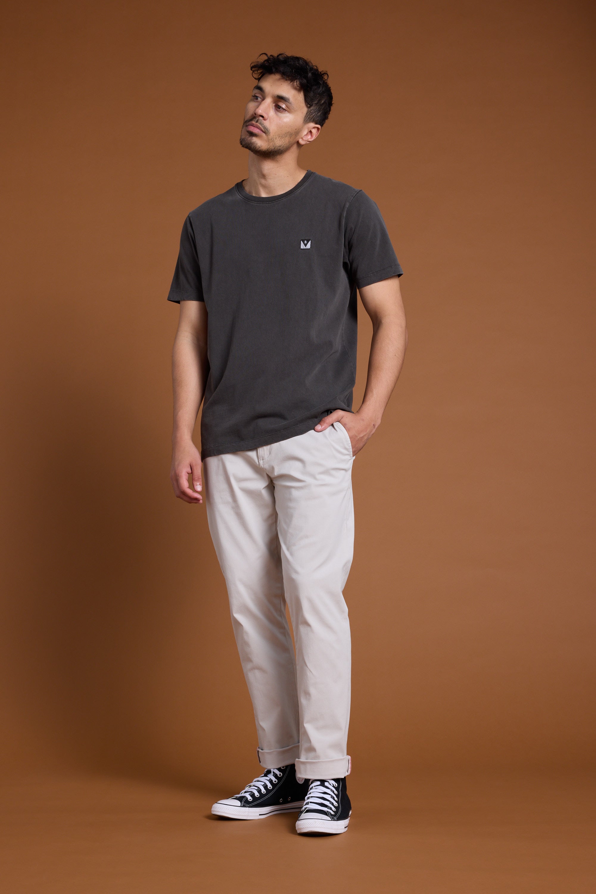 A man with short dark hair stands against a brown background, wearing the Barkers Newton Washed Tee in dark grey, paired with light beige pants and black high-top sneakers. His left hand is in his pocket as he looks slightly upwards.