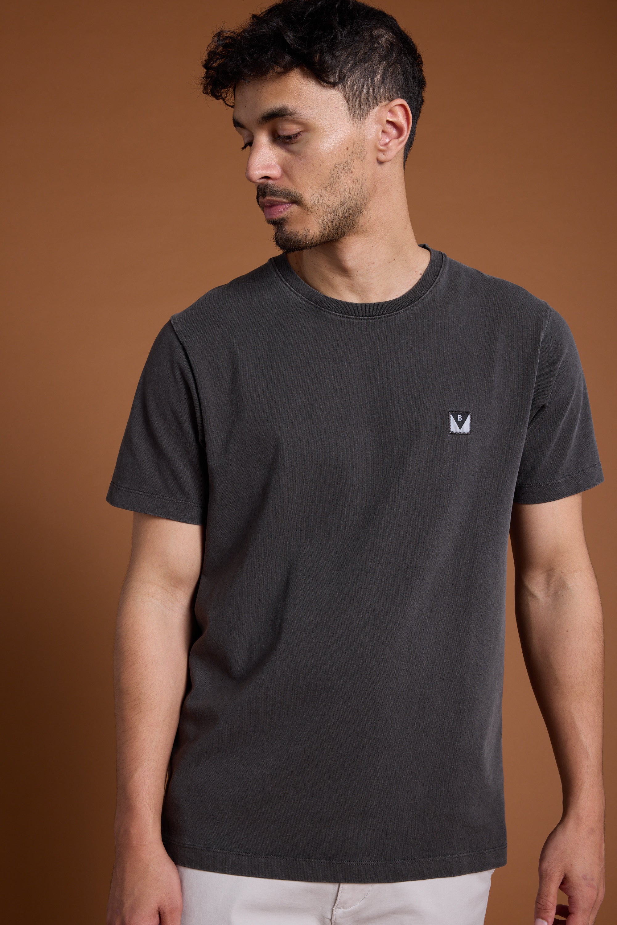 A man with short, dark curly hair and a trimmed beard models the Barkers Newton Washed Tee, a faded charcoal shirt with a small white chest logo, against a plain brown background, looking down to his left.